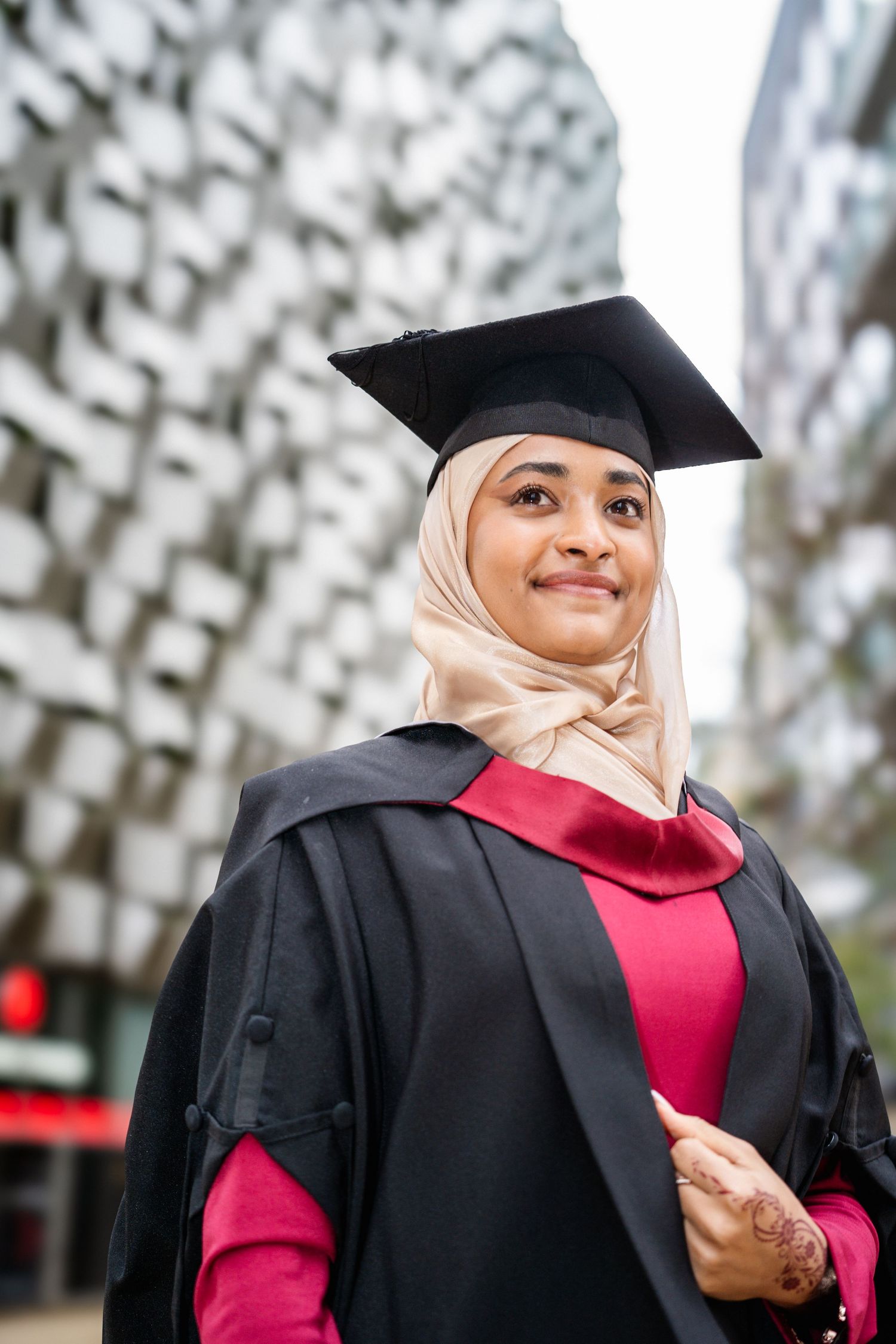 Graduate in black cap and gown with red dress smiles joyfully in front of blurred background.