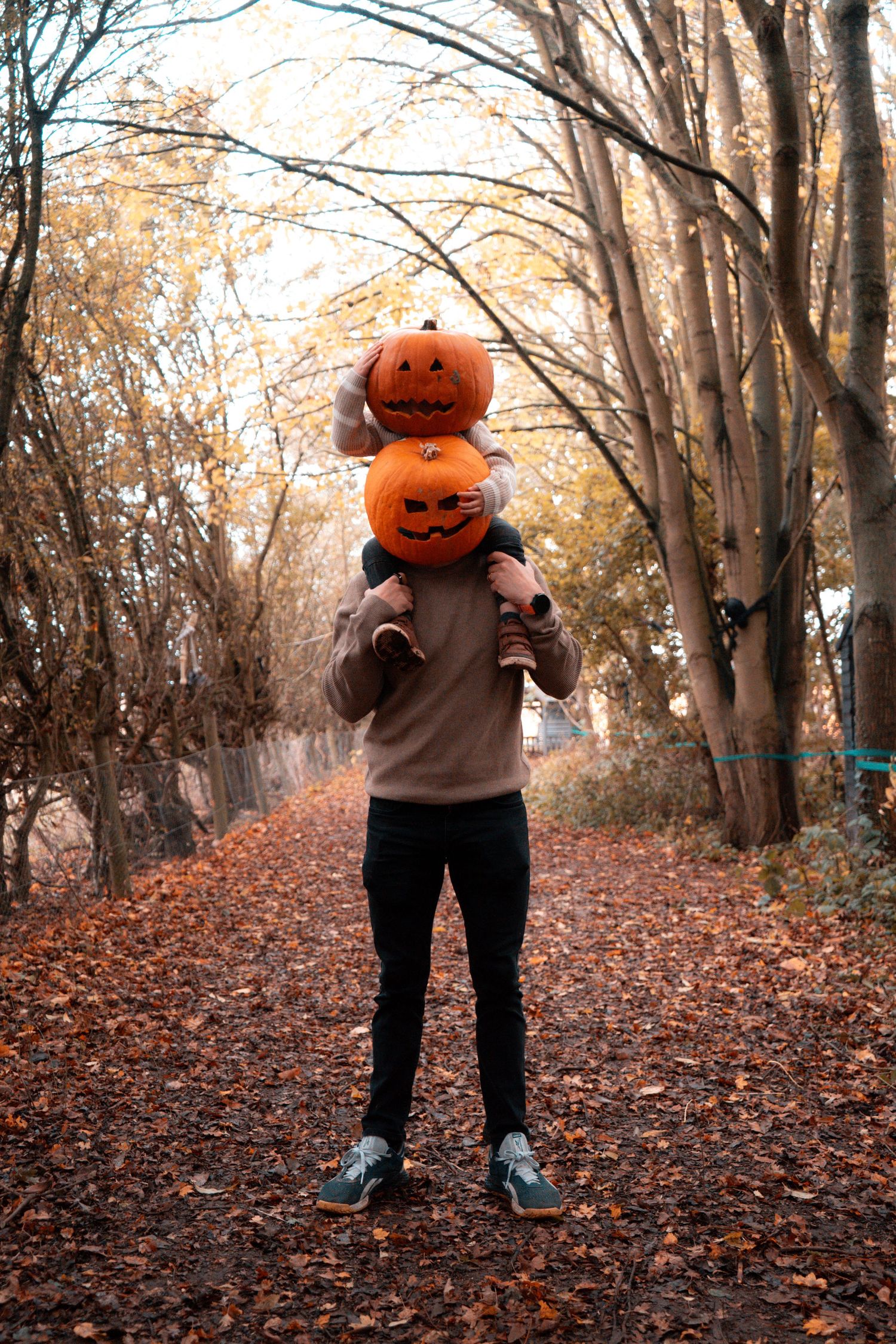 Figure wearing pumpkin head walks through forest path covered in fallen autumn leaves.