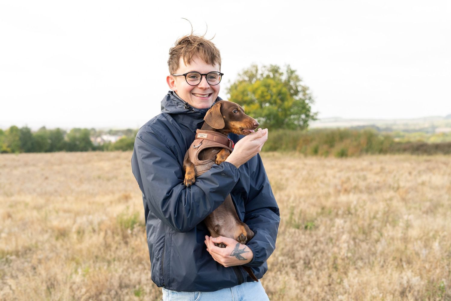 A person in a navy jacket holding and cuddling a small brown dachshund puppy in a field.