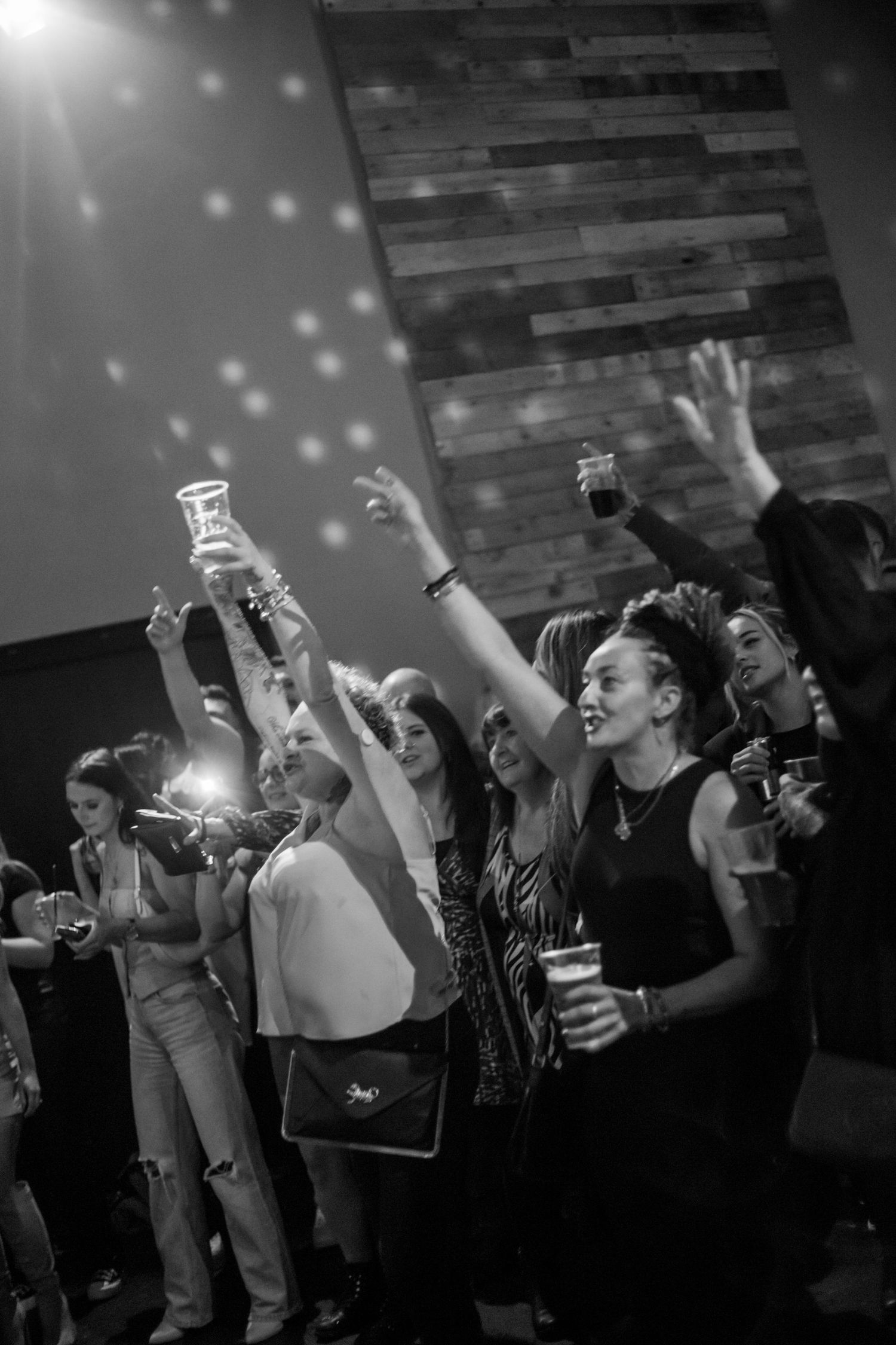 Concert audience raises drinks and hands during performance under disco lights.