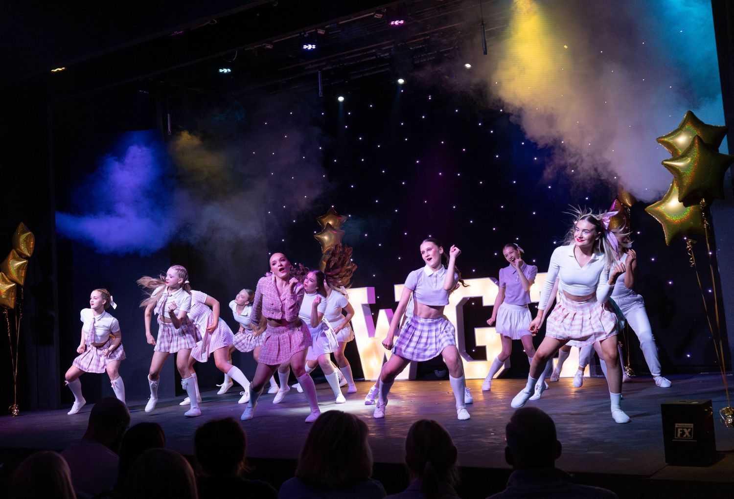 Dance ensemble performs in coordinated pink and white outfits under dramatic stage lighting.