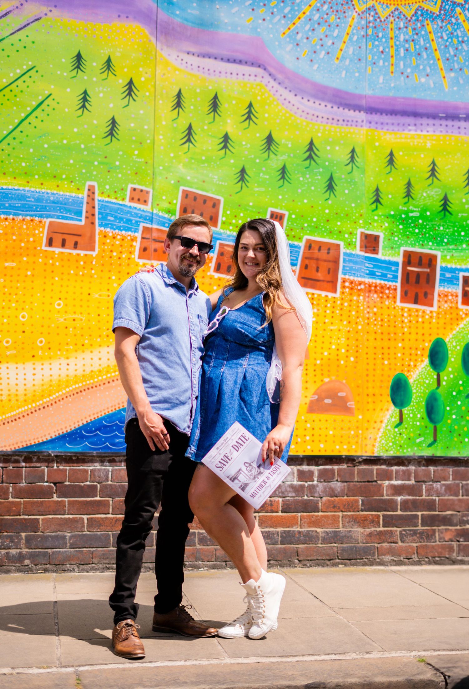 Couple sitting by whimsical mural depicting colorful landscape with houses and trees.