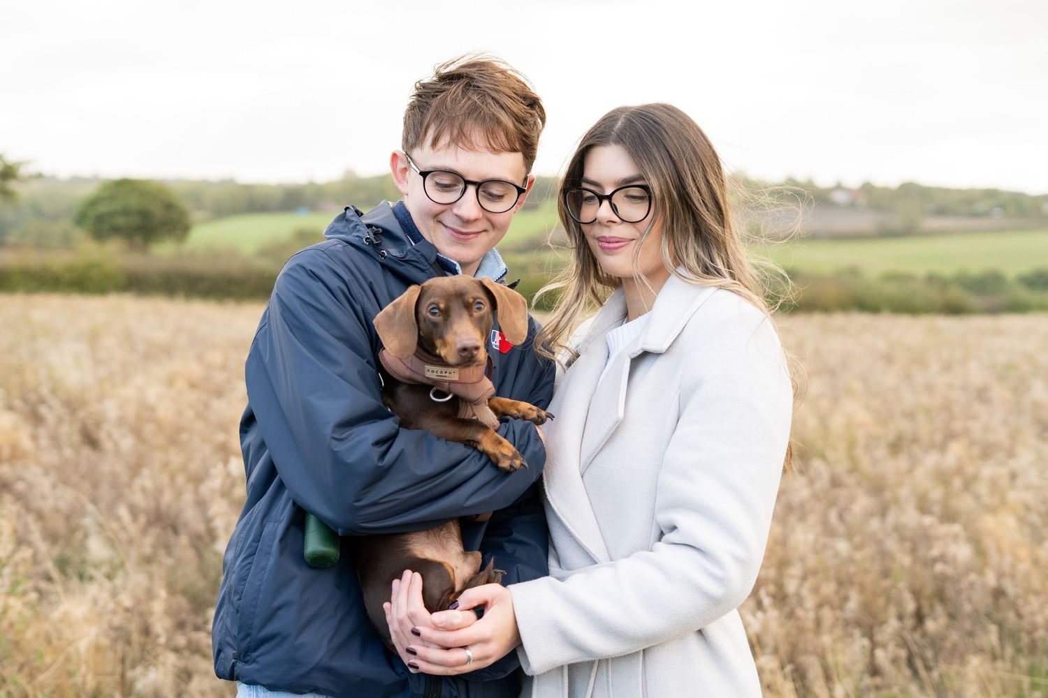 A couple embracing their dachshund puppy in a rustic field setting.