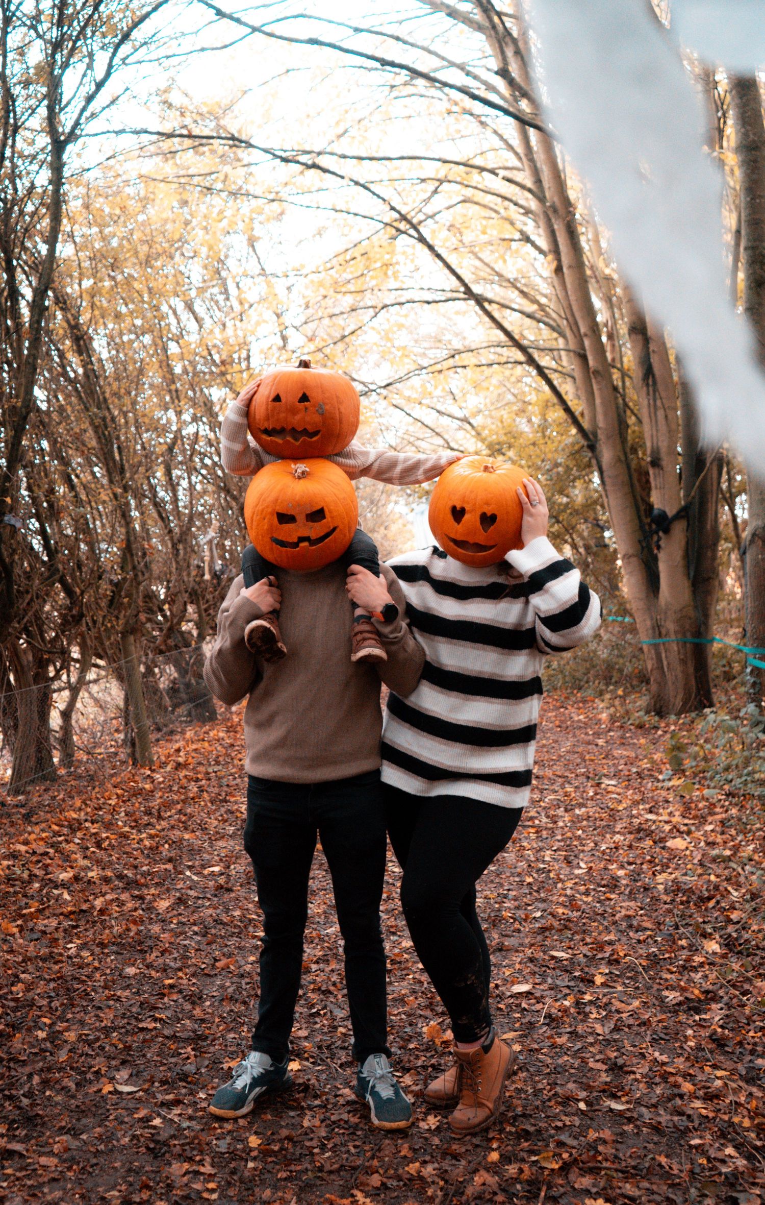 Two figures with pumpkin heads stand together on a leaf-covered autumn forest trail.