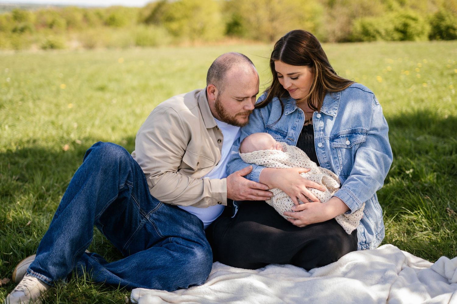 Family lying together on blanket in sunny field with newborn baby.