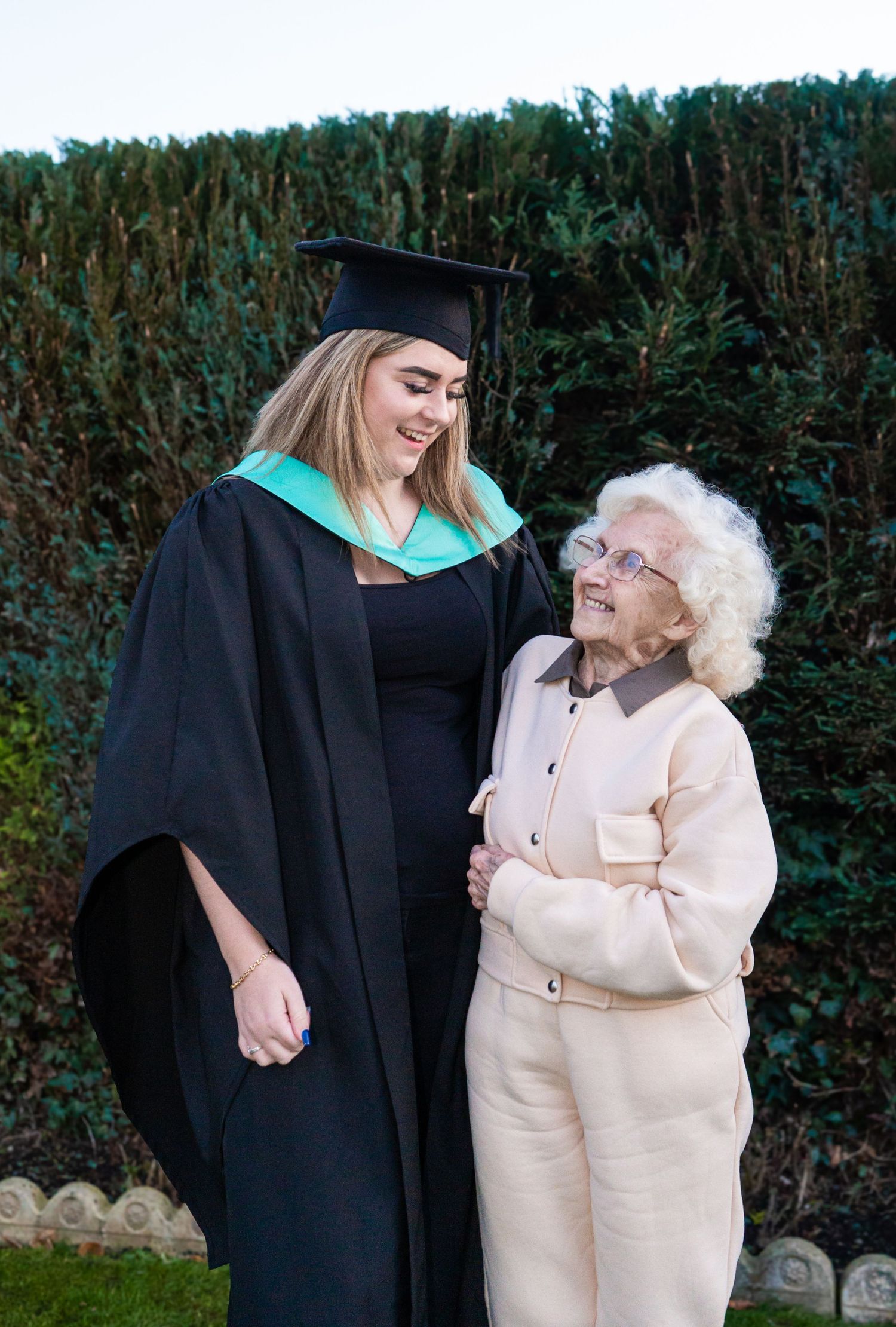 Graduation celebration photos with graduate in black gown sharing laughs with elderly relative by hedge.