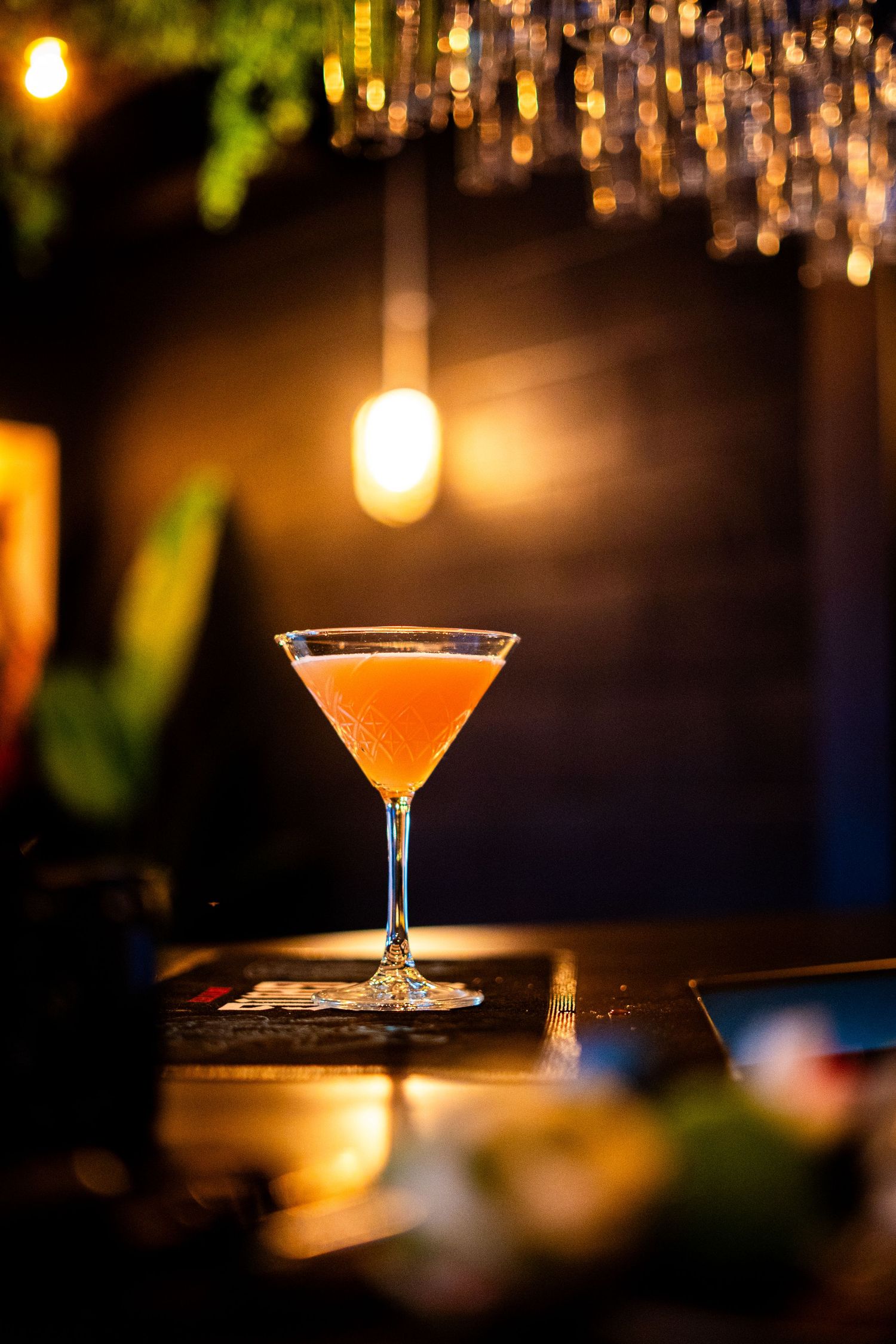 Orange-hued cocktail in coupe glass under warm pendant lighting at bar.
