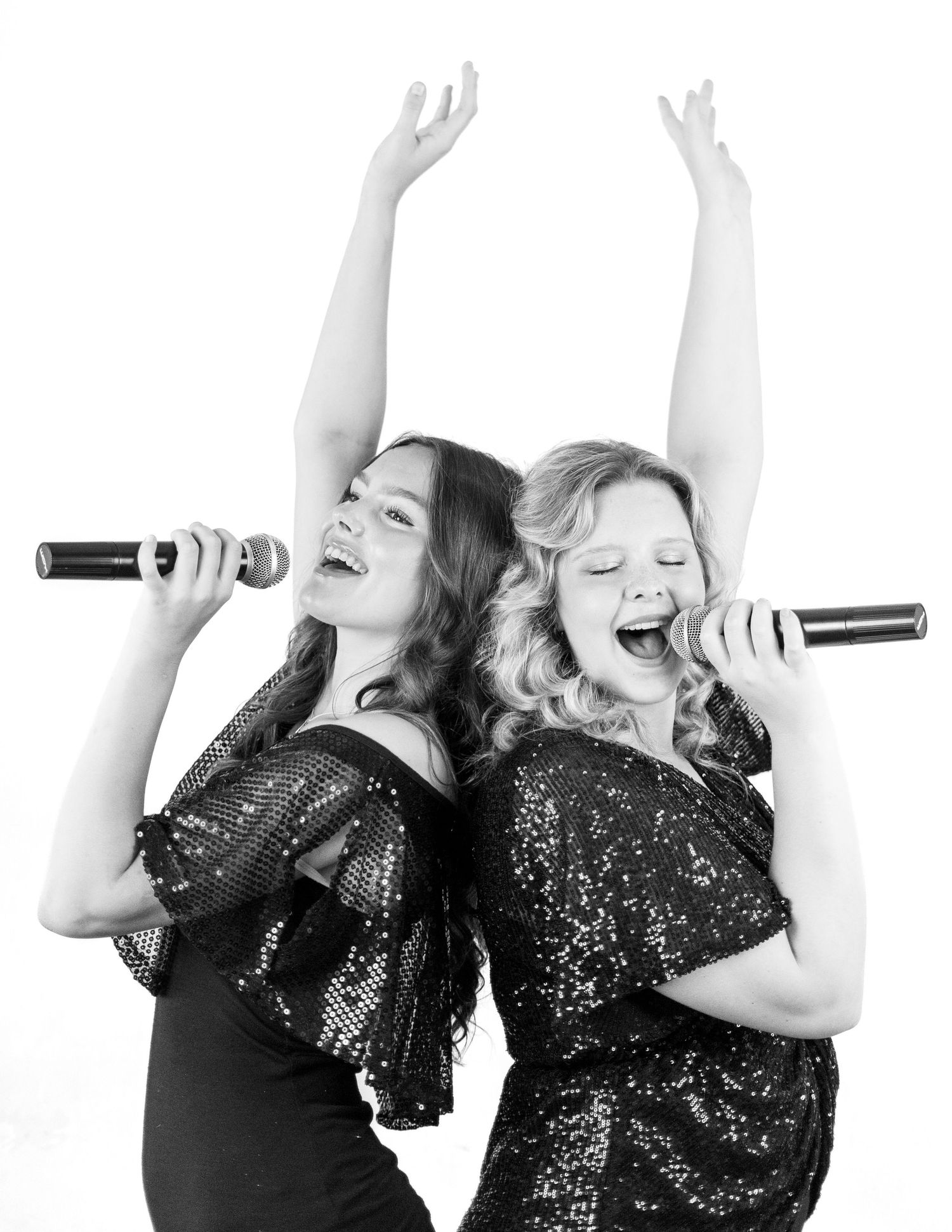 Two performers in black sparkly outfits sing and dance energetically against white backdrop.