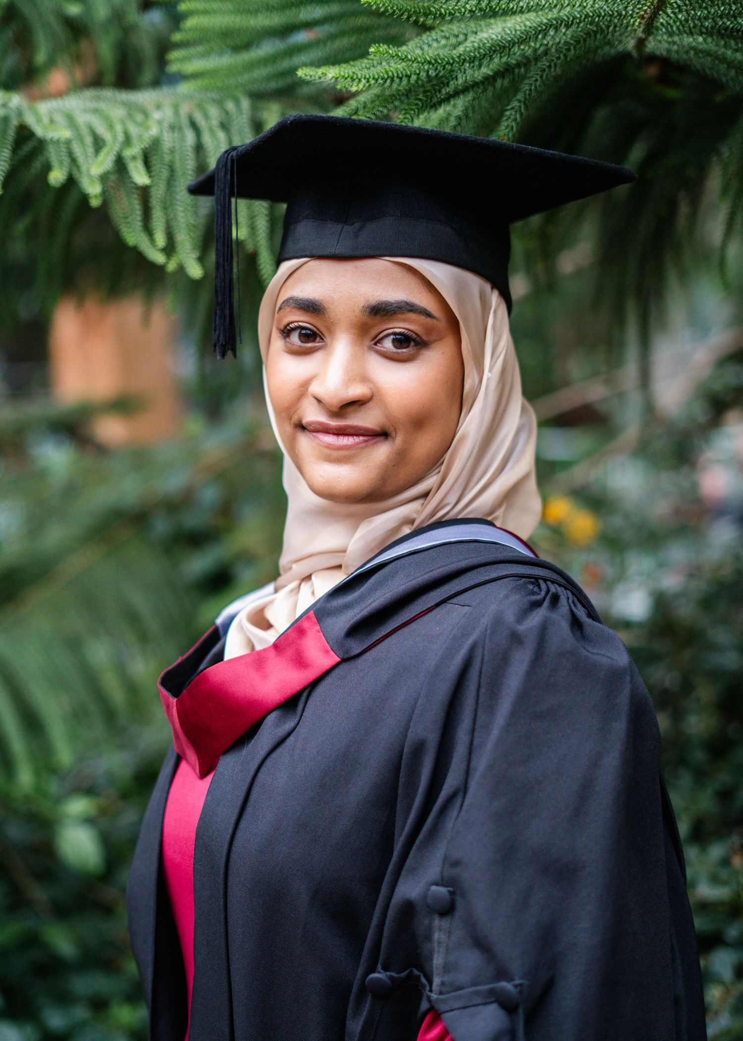 Graduate in academic regalia poses against lush green foliage background.