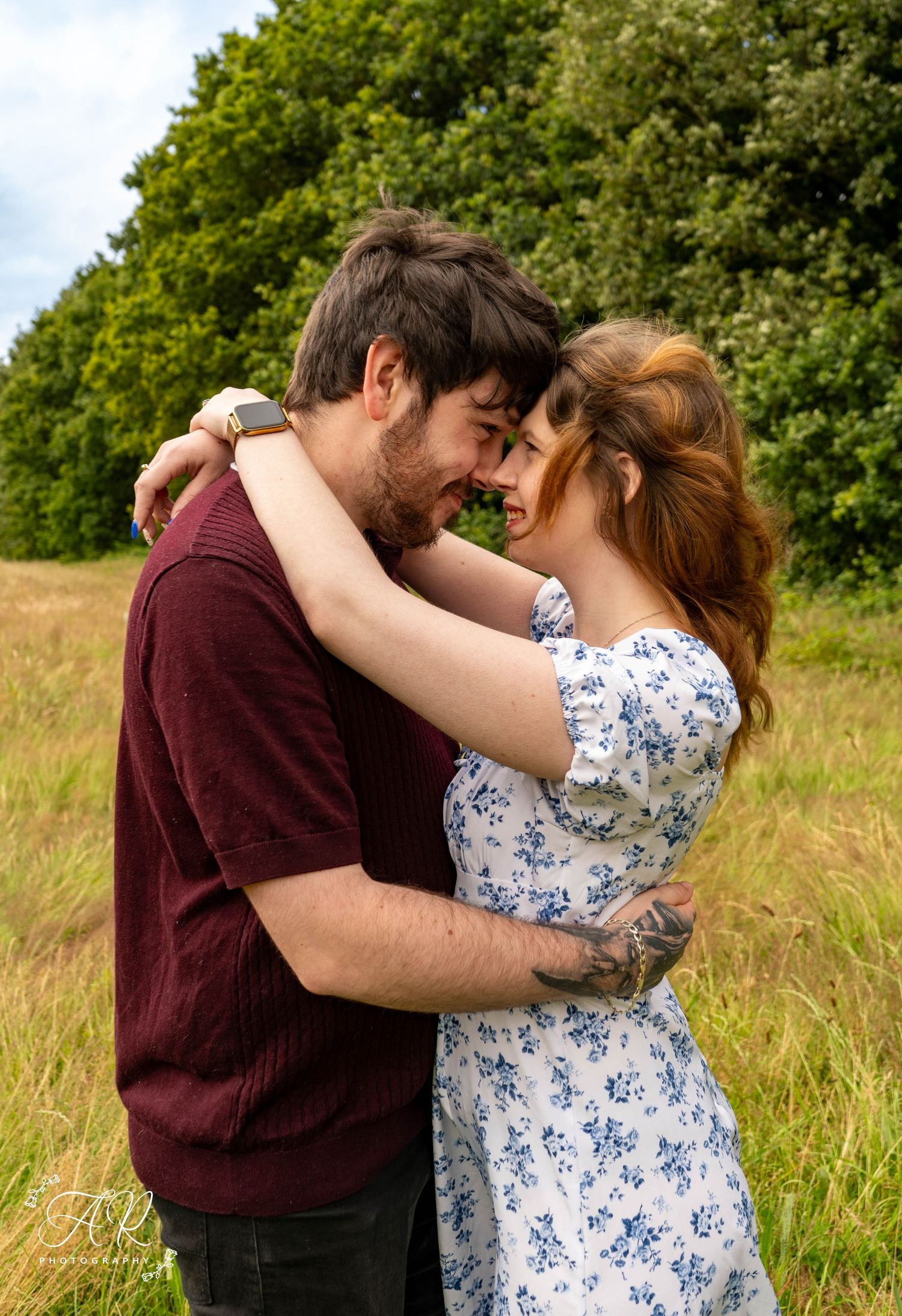 A romantic couple shares tender moments in a field with green trees in the background.