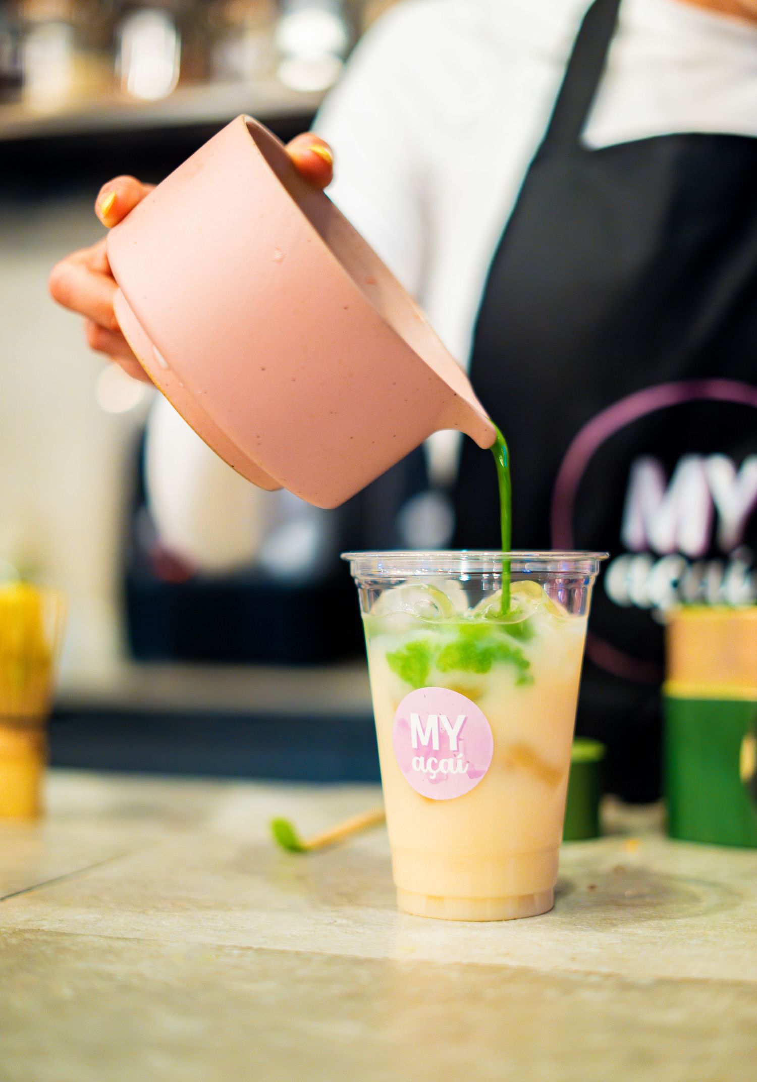 Pink teapot pouring green liquid into branded beverage cups.