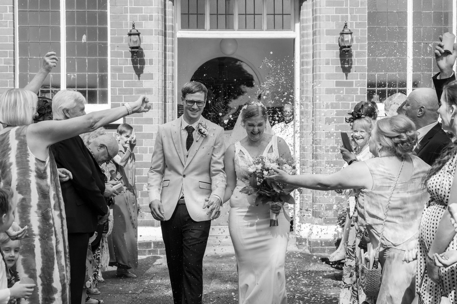 Wedding party celebrates with confetti outside a traditional brick building.