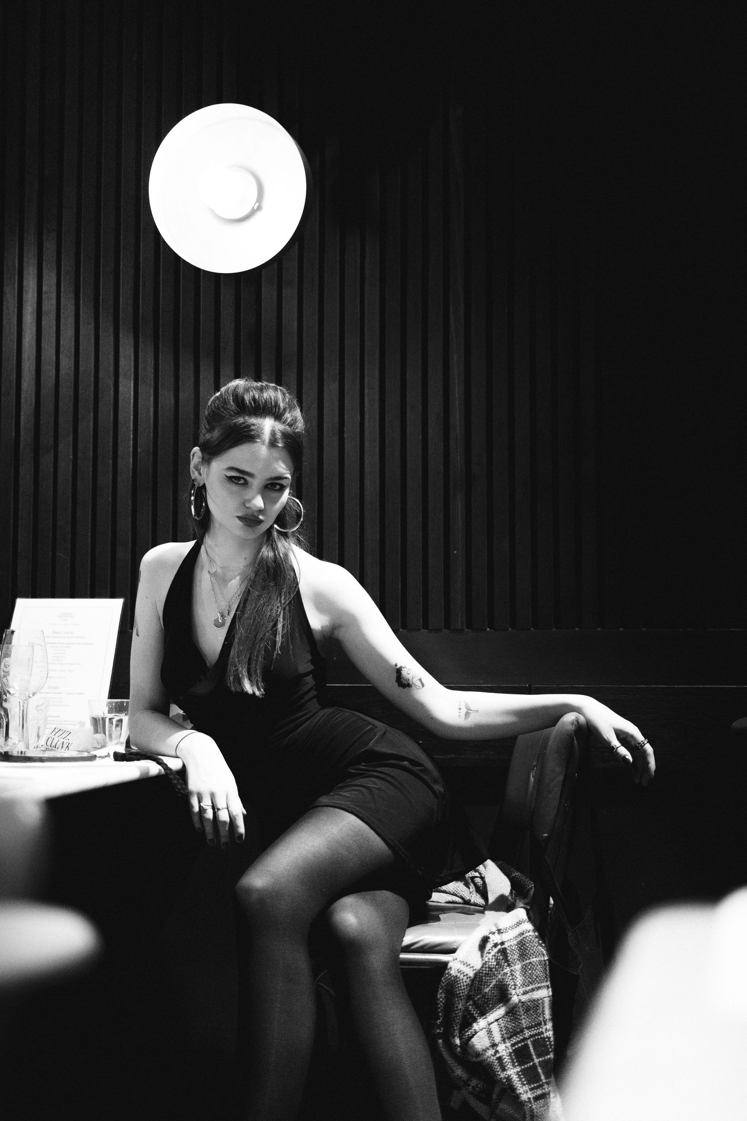 A black and white photo of someone in elegant attire seated at a bar under a circular light.