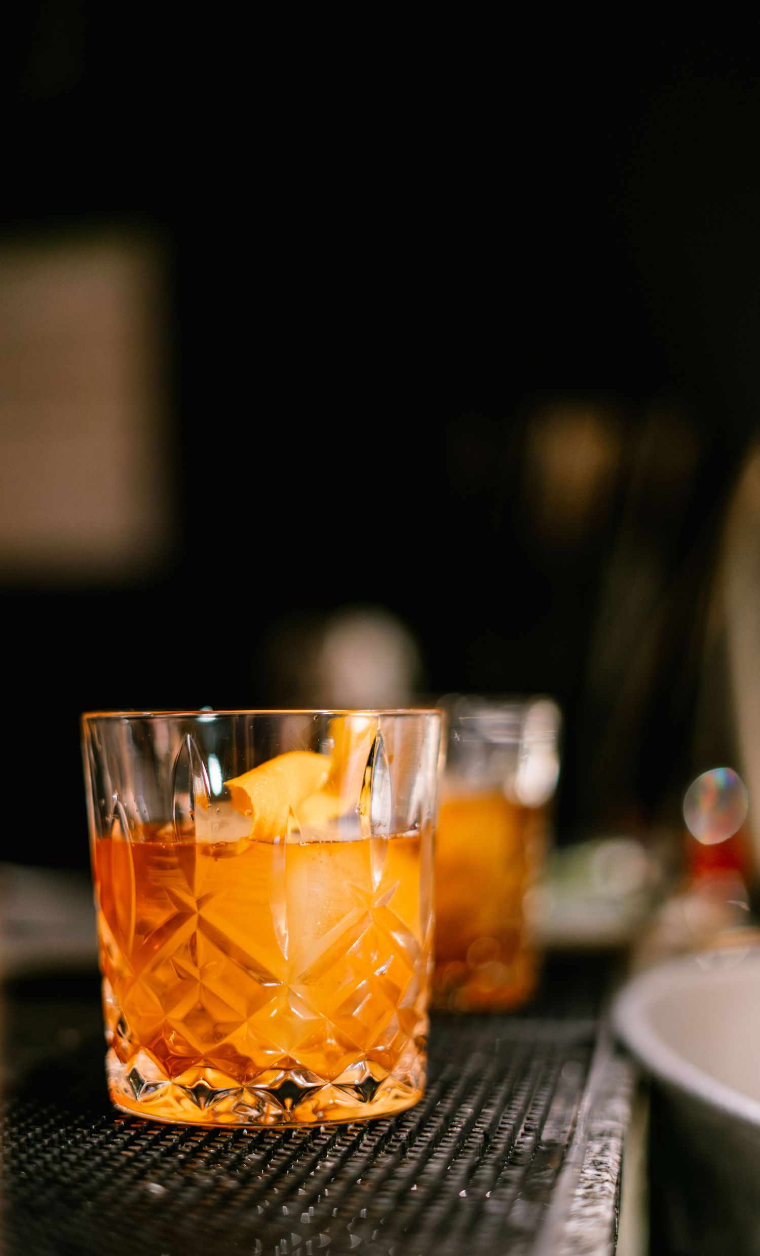 Crystal cocktail glasses with amber-colored drinks lined up on a black bar mat.
