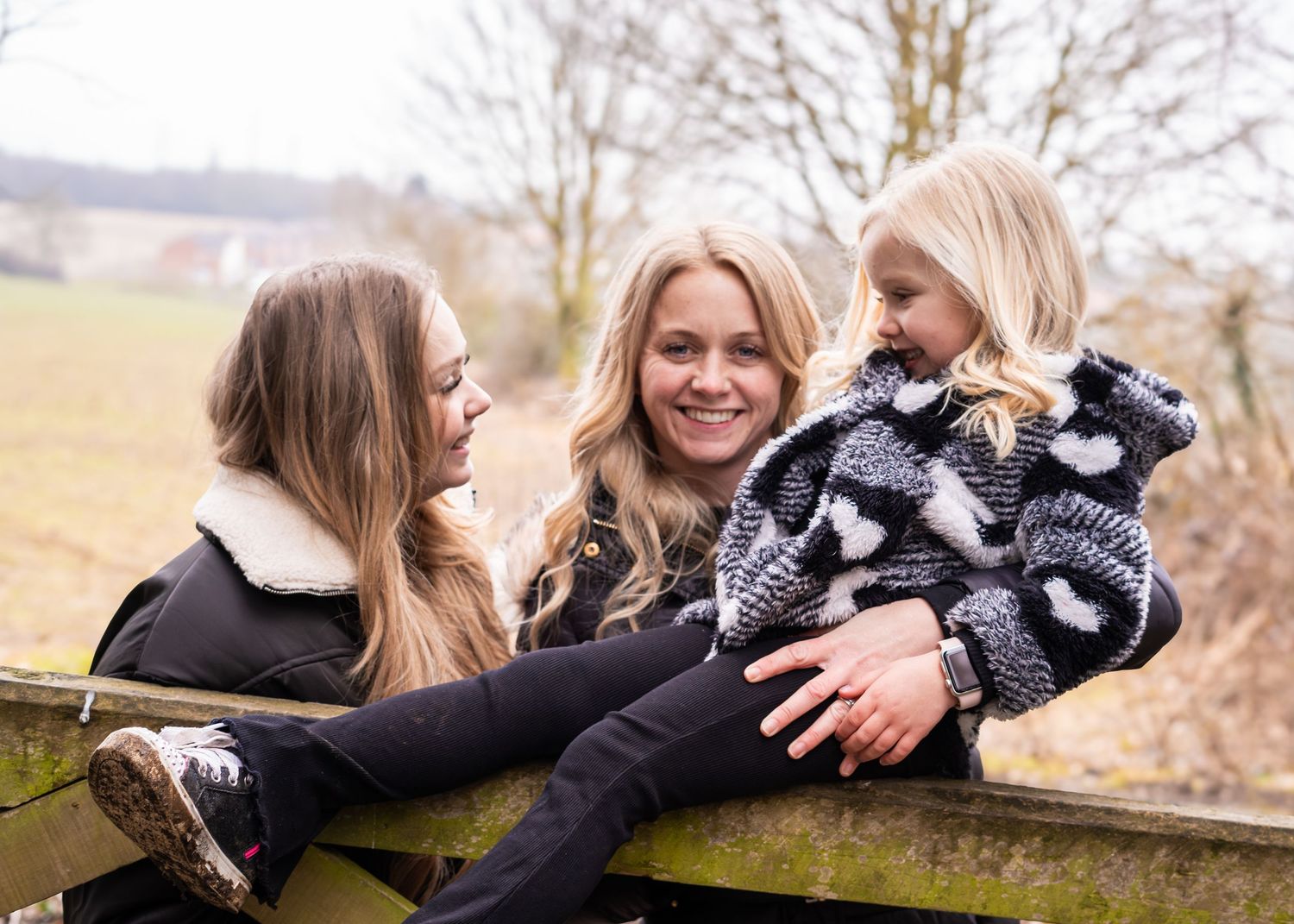 Three people laugh together while relaxing on a wooden gate in the countryside.