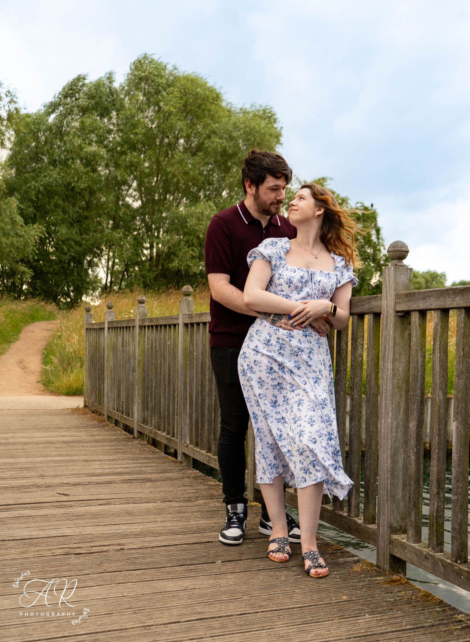 Loving couple poses on wooden boardwalk with natural scenery in background.