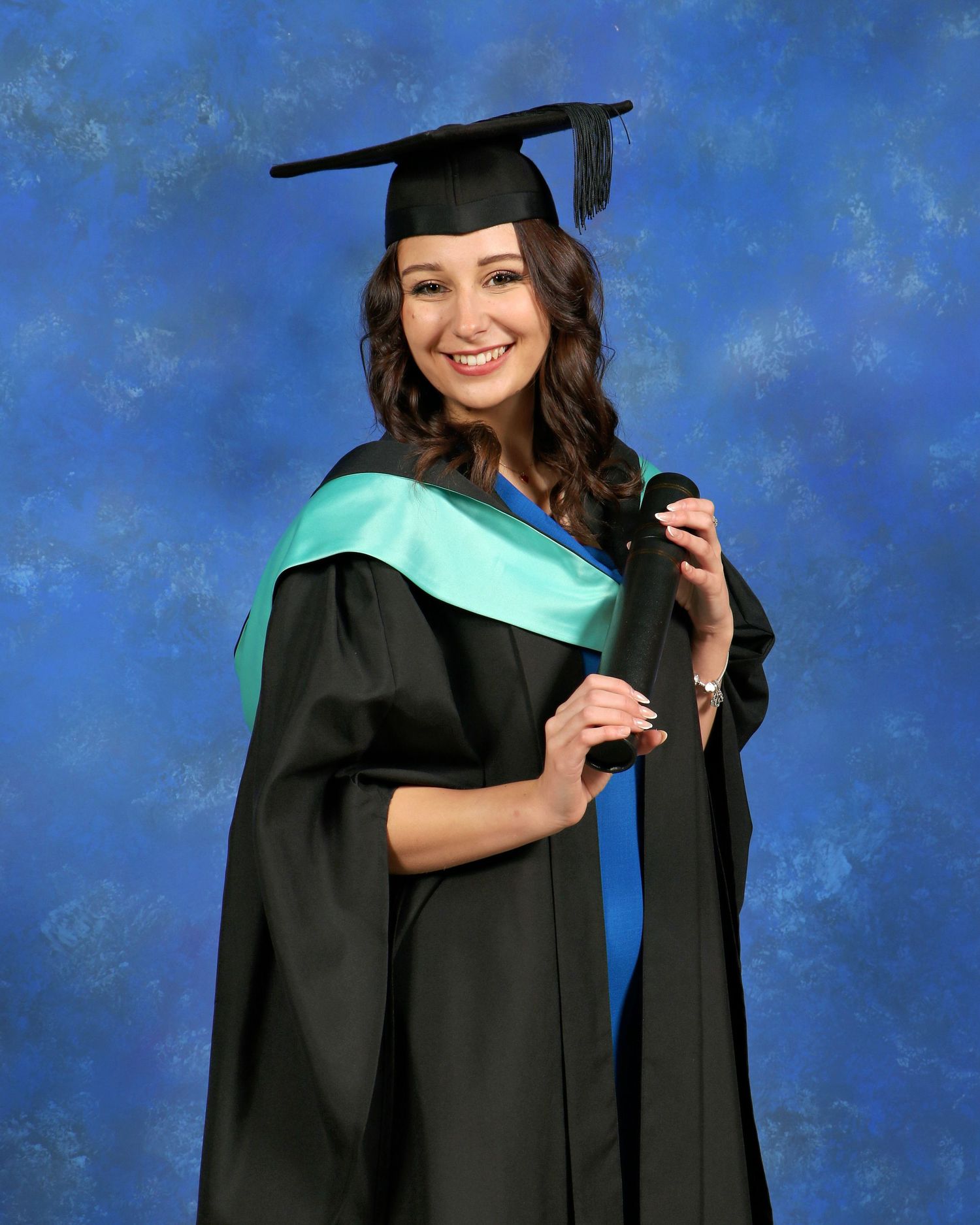 A graduate in cap and gown with teal stole poses against blue backdrop.