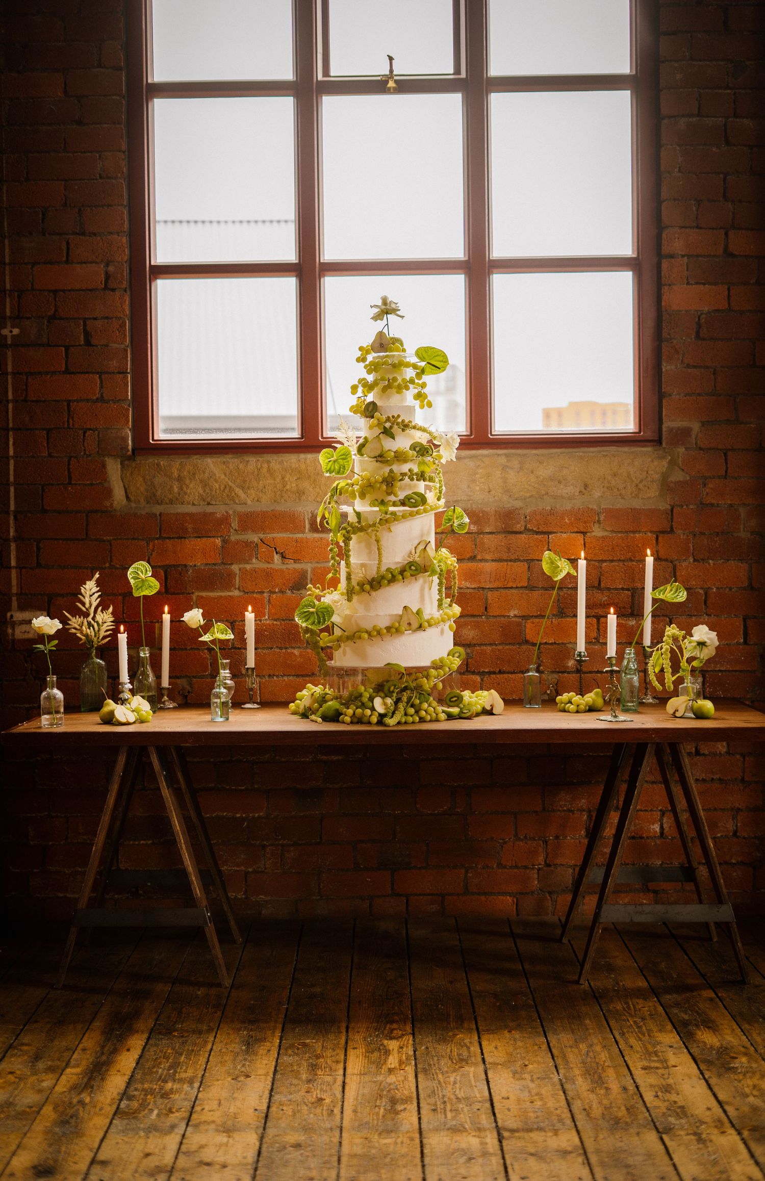 Wedding cake display with candles and greenery in a rustic brick-walled venue with wooden floors.