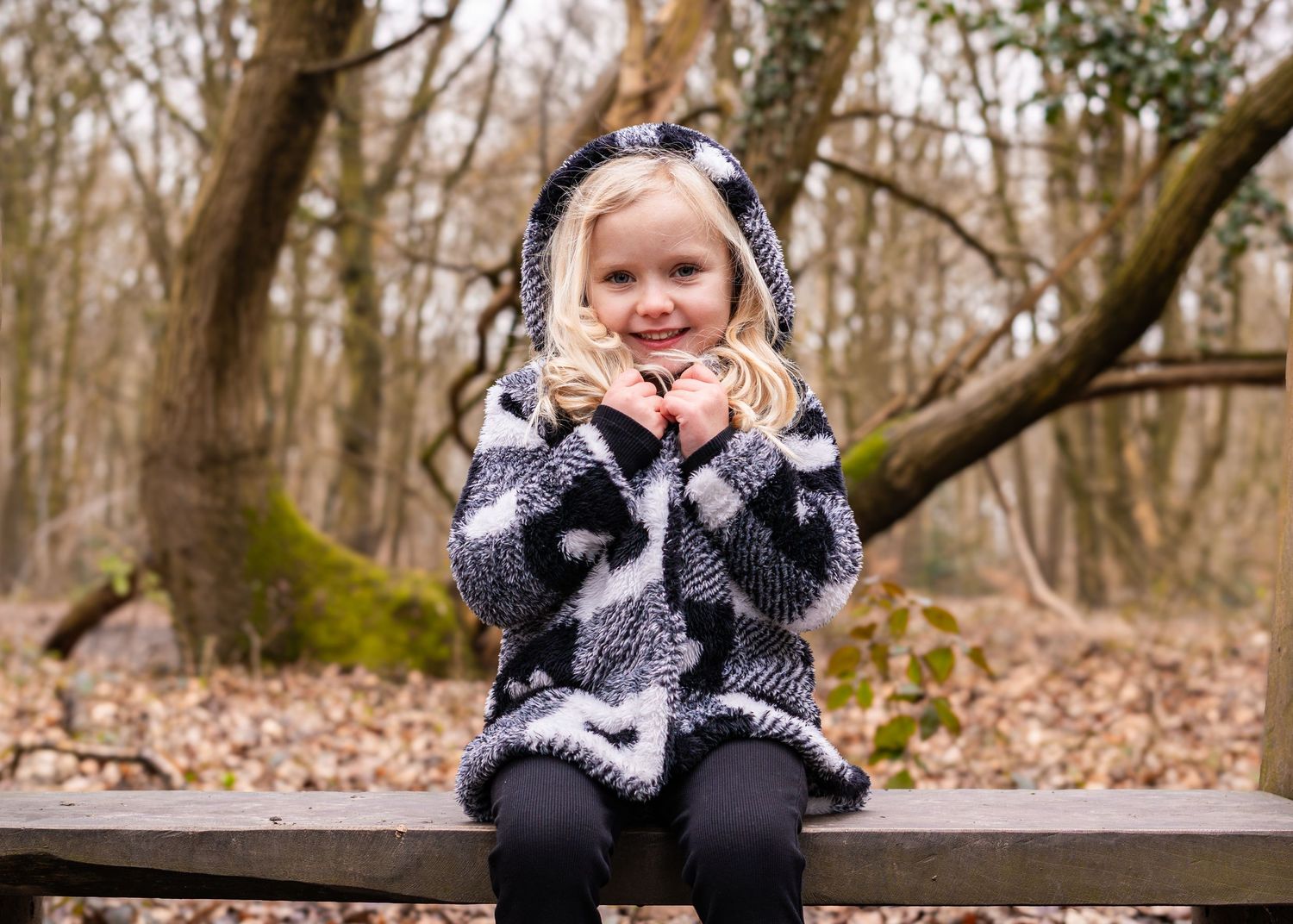 A child in winter clothing sits on a wooden bench surrounded by fallen leaves.