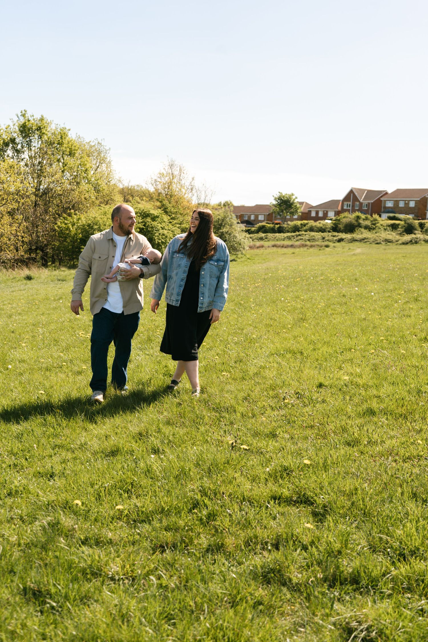 Couple walking together through a sunny green field with houses in background.