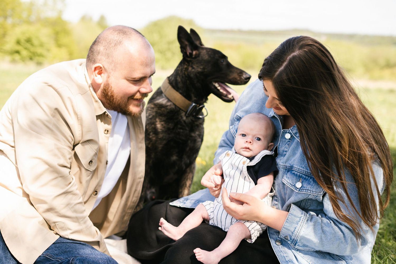 Family and black dog relaxing together on blanket in grassy field on sunny day.
