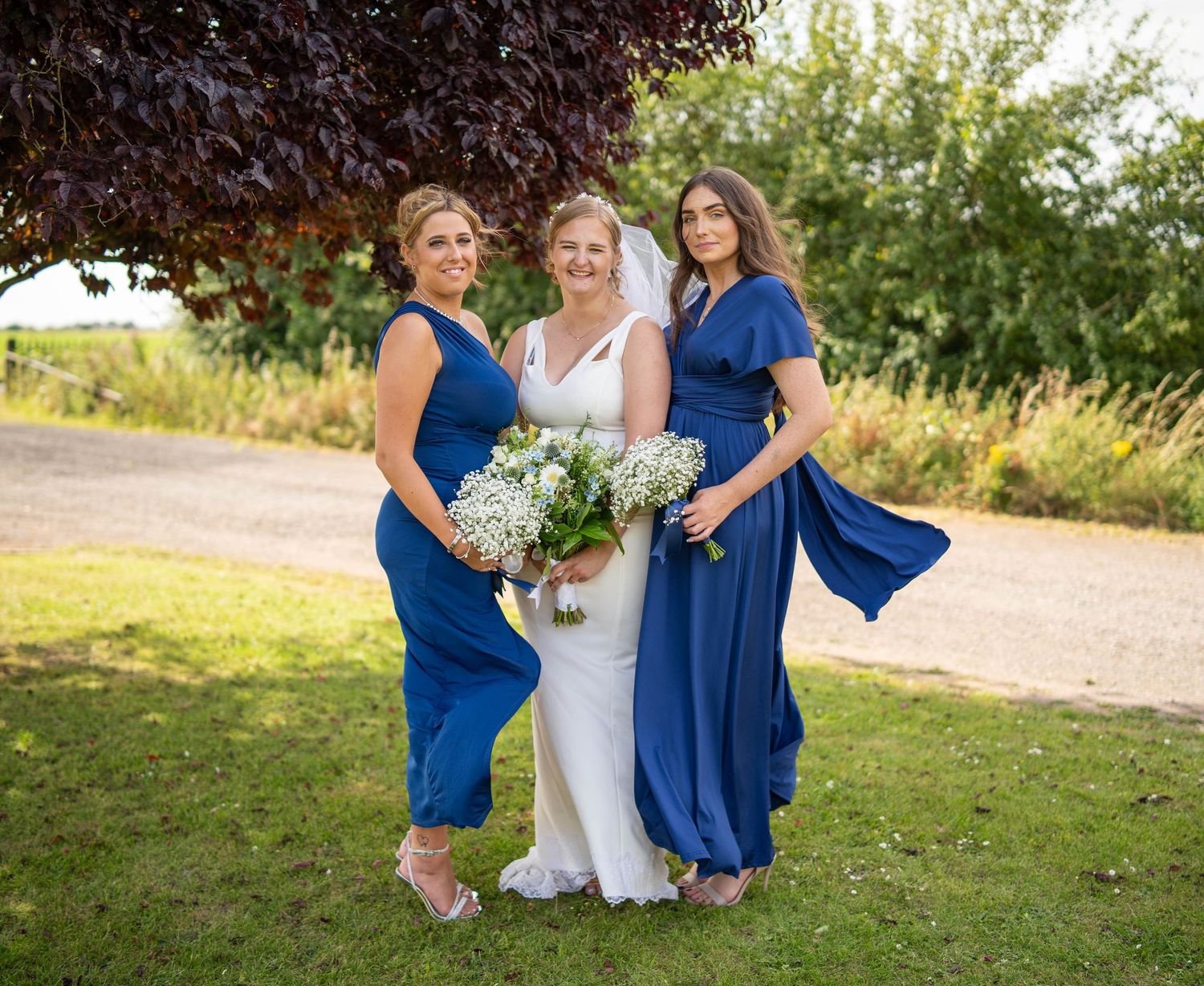 Bridal party in navy blue dresses poses outdoors with bouquets on a sunny day.