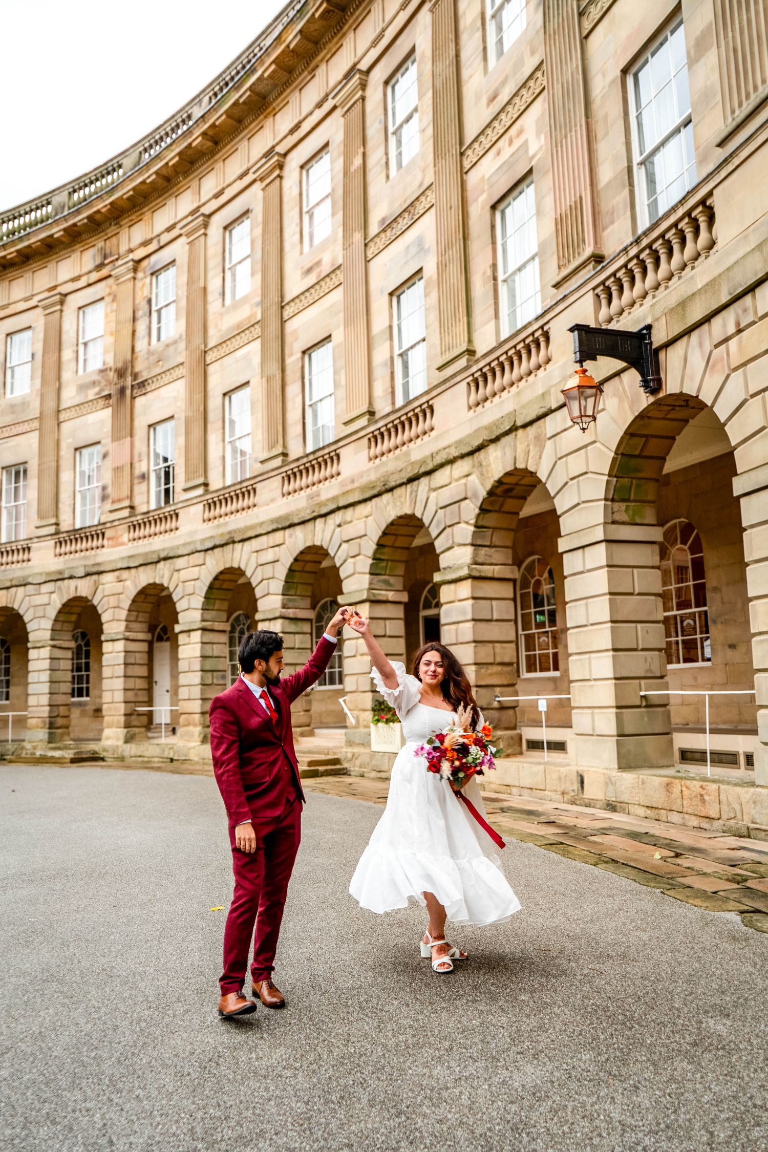 A couple celebrates joyfully in front of a historic stone building with arched architecture.