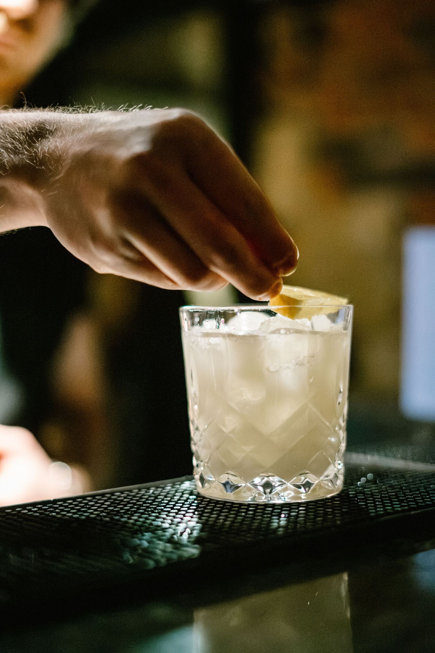 Bartender pouring a white cocktail over ice in a crystal glass on a black bar mat.