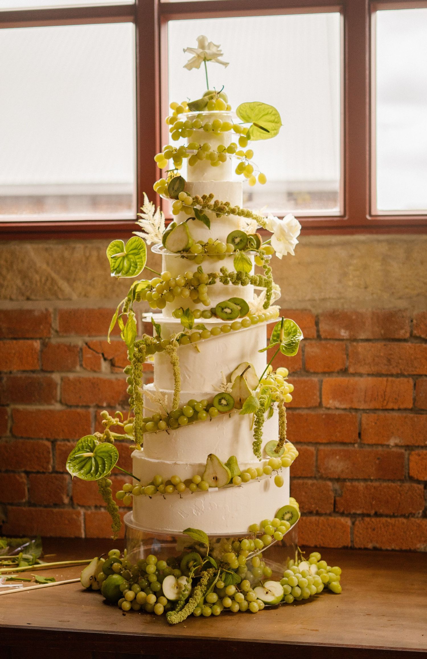Multi-tiered white cake decorated with spiraling green grapes and leaves in an industrial loft setting.