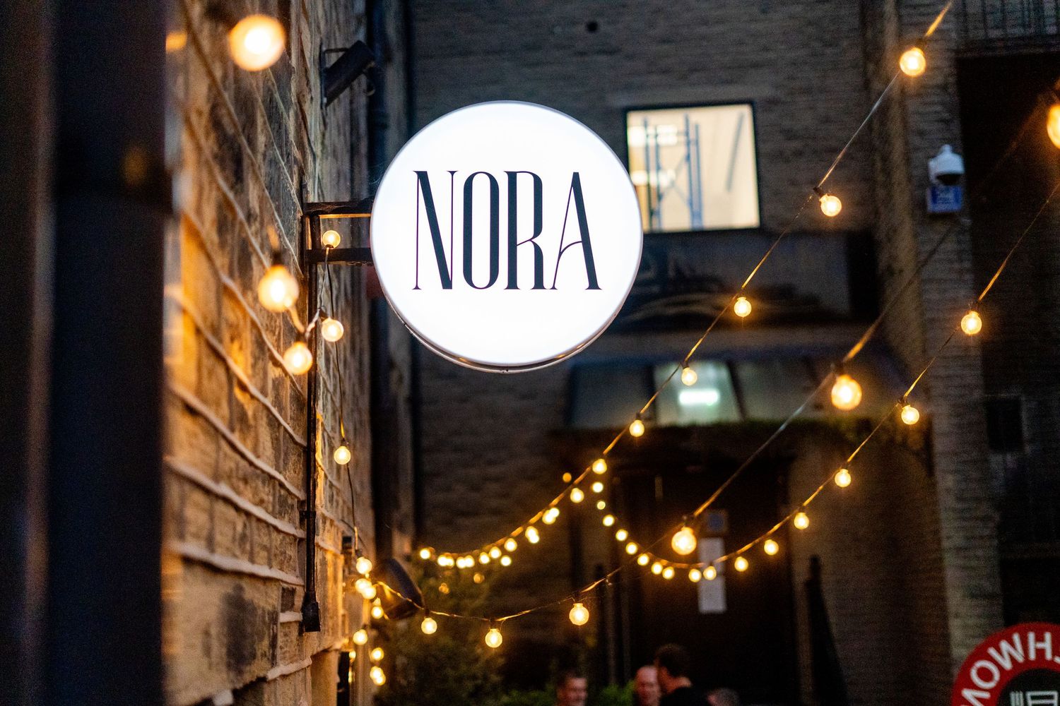 Round white NORA sign mounted on brick wall with decorative string lights.