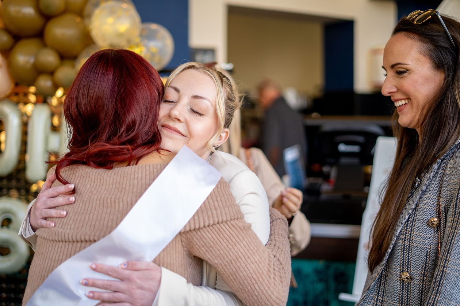 Friends sharing joyful embraces at a celebration with balloon decorations in the background.
