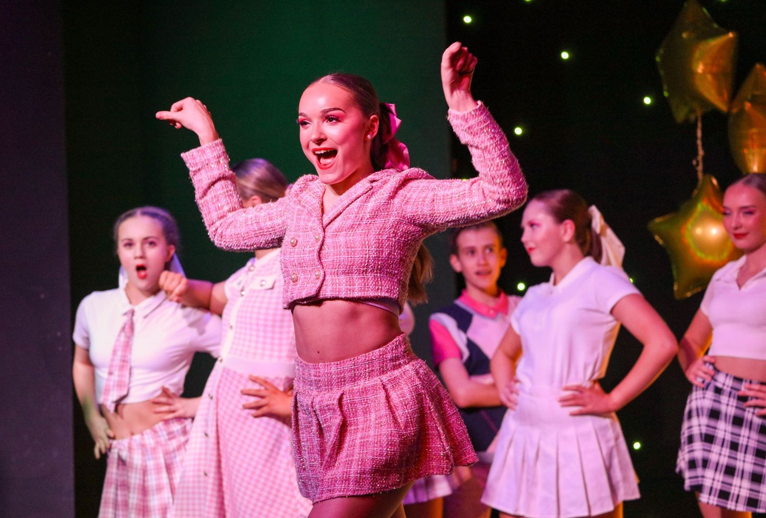 Performers in pink outfits dance energetically on stage during a theatrical production.