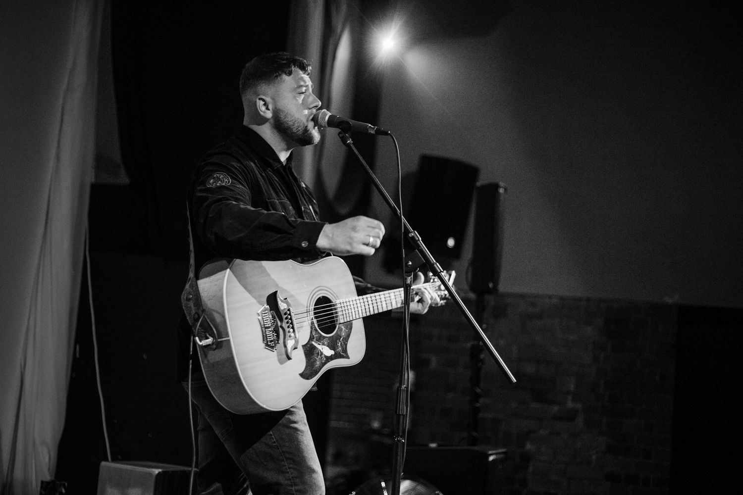Dramatic black and white performance shot of a musician playing acoustic guitar on stage.
