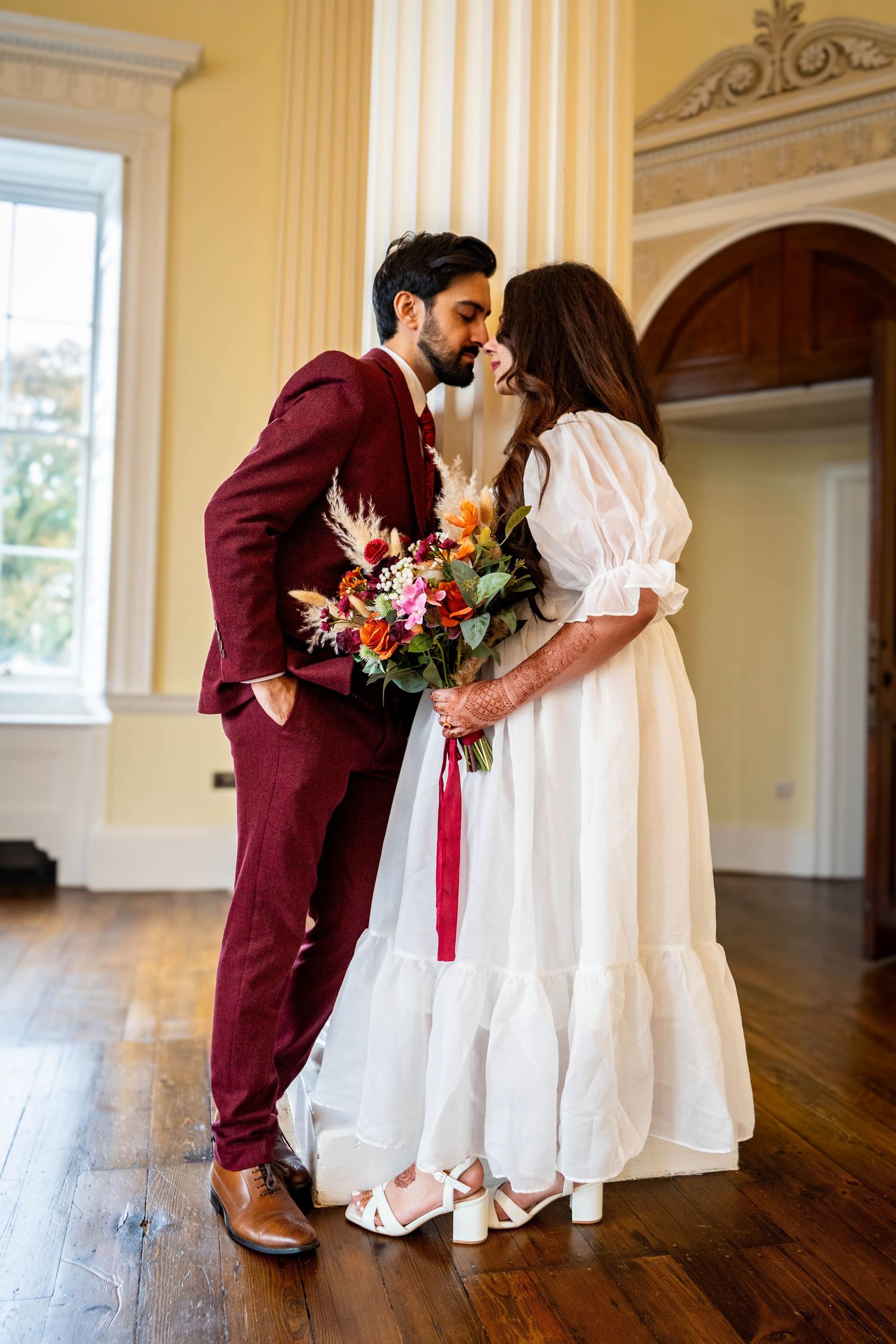 Wedding couple in burgundy and white attire posing in an elegant yellow-walled room.