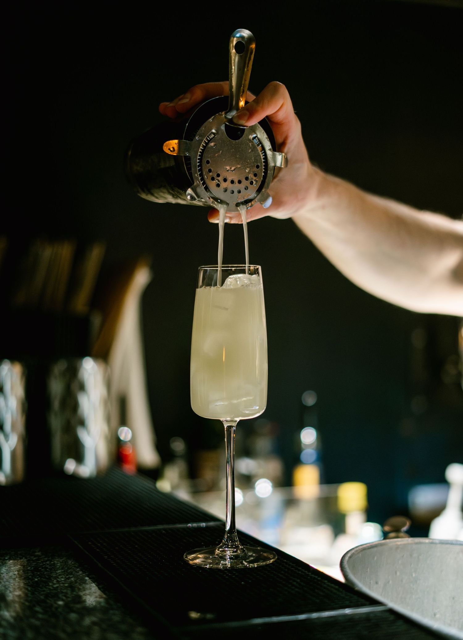 Bartender straining a light-colored cocktail into a glass with ice.