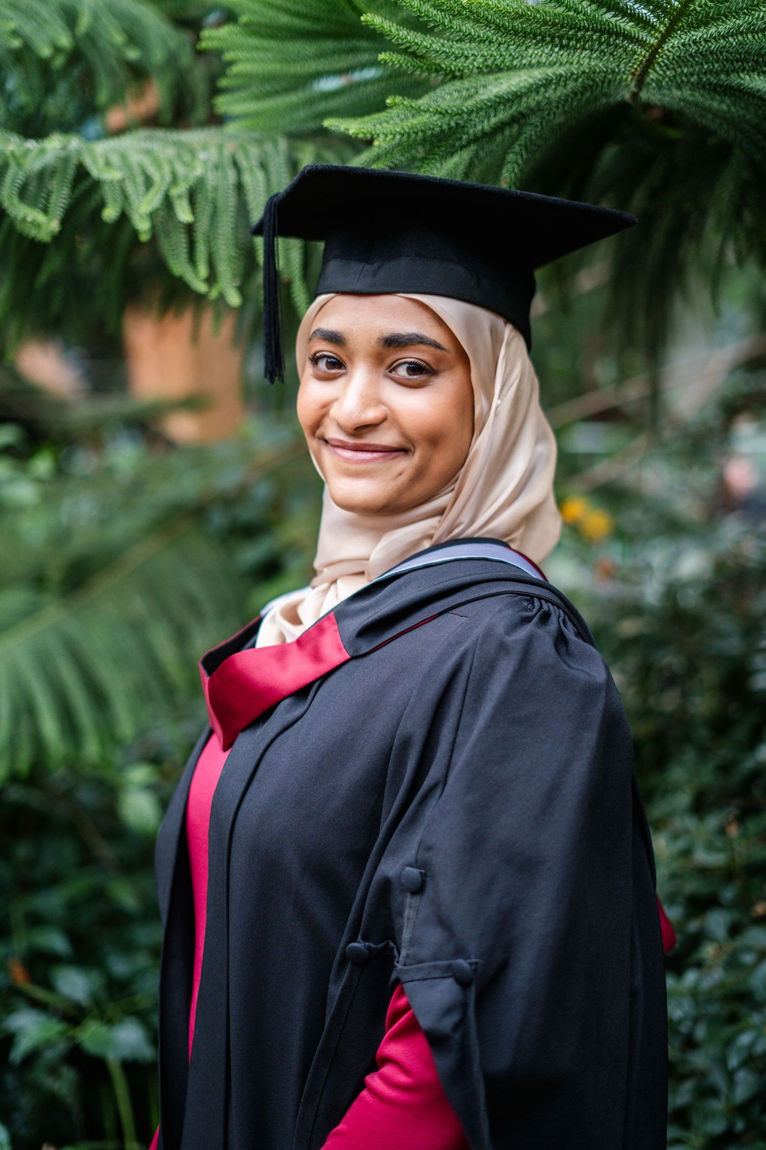 Graduate in black cap and gown poses with greenery in background.