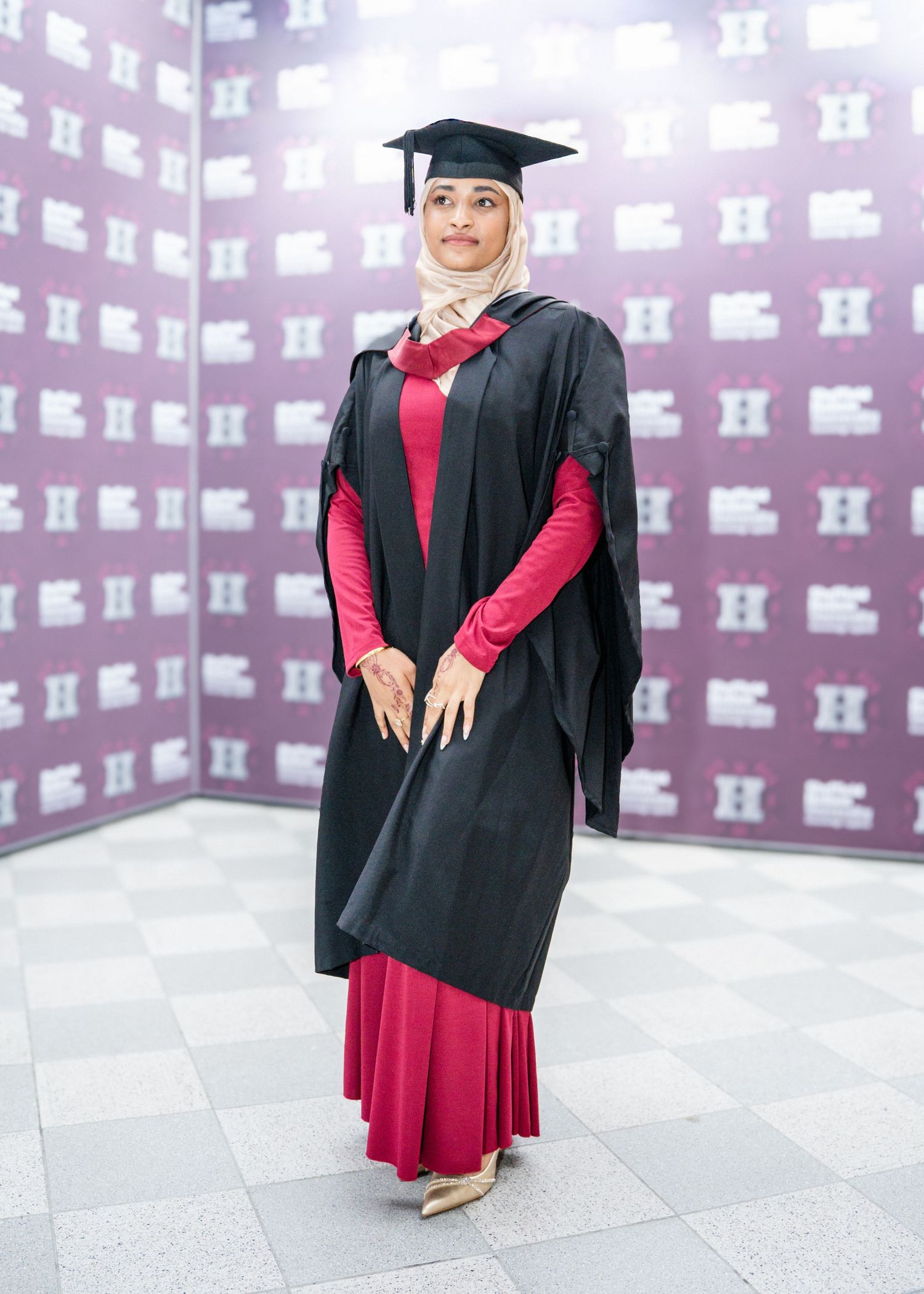 Graduate in black cap and gown with red dress poses against purple and white backdrop.