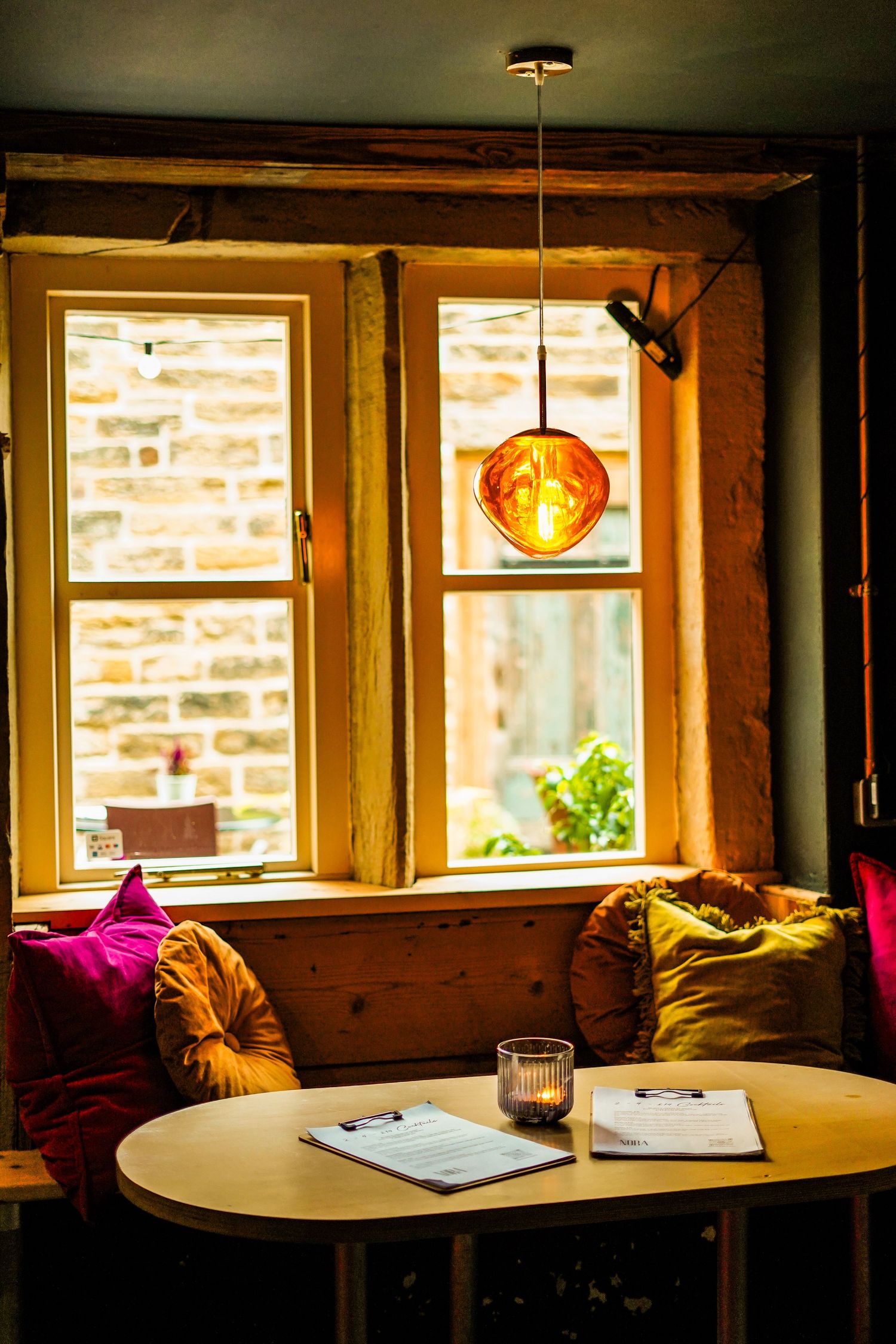 Cozy cafe corner with round wooden table, colorful cushions and pendant light by sunlit windows.