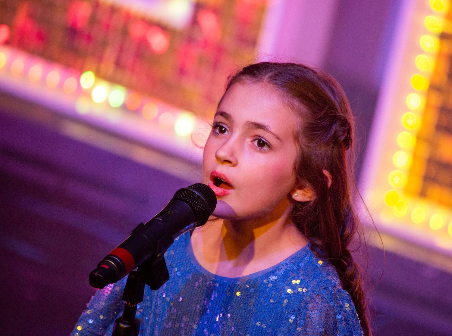 Performer in sparkly blue dress sings into microphone under purple stage lighting.