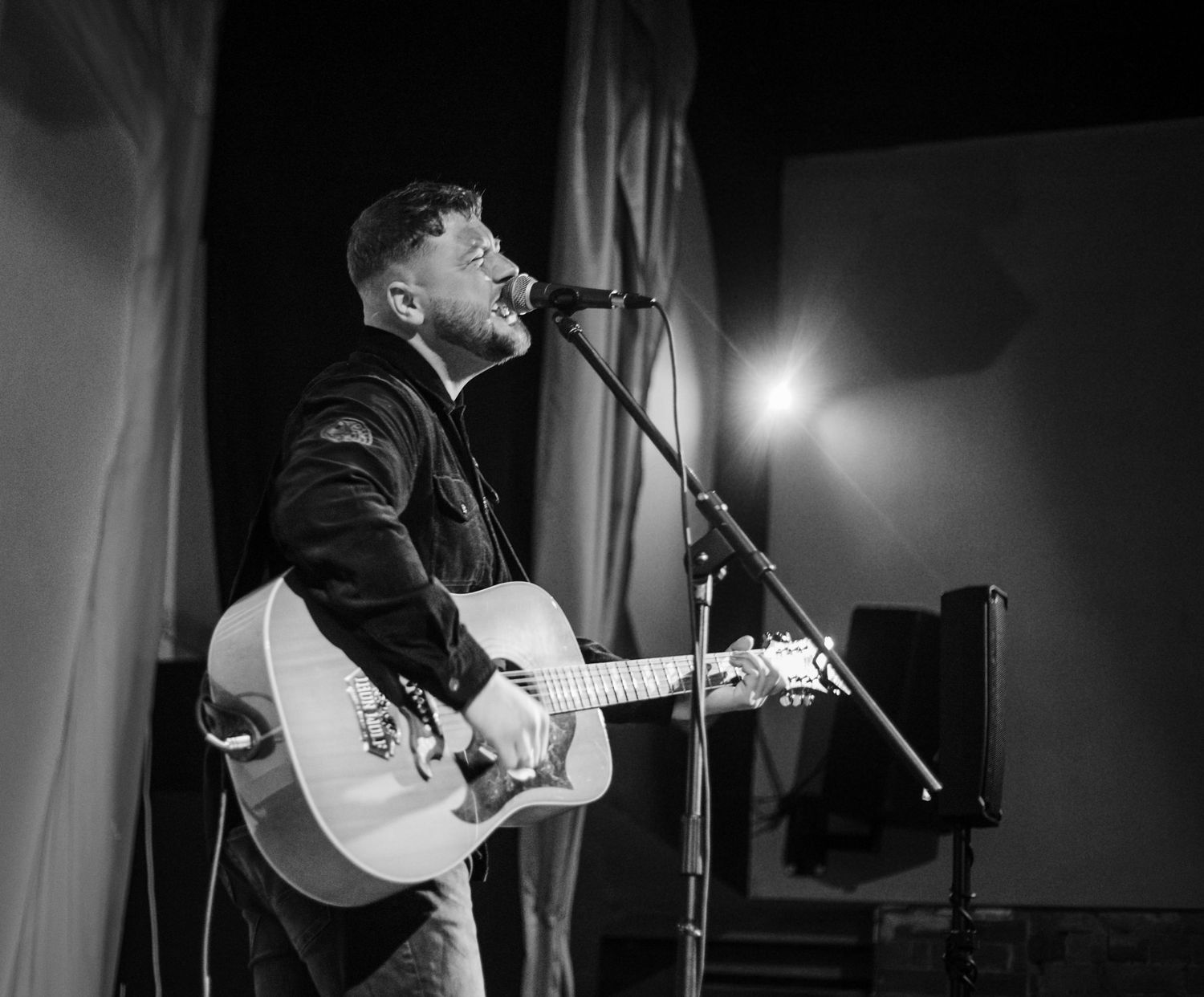 Black and white photo of a performer on stage with an acoustic guitar under dramatic lighting.