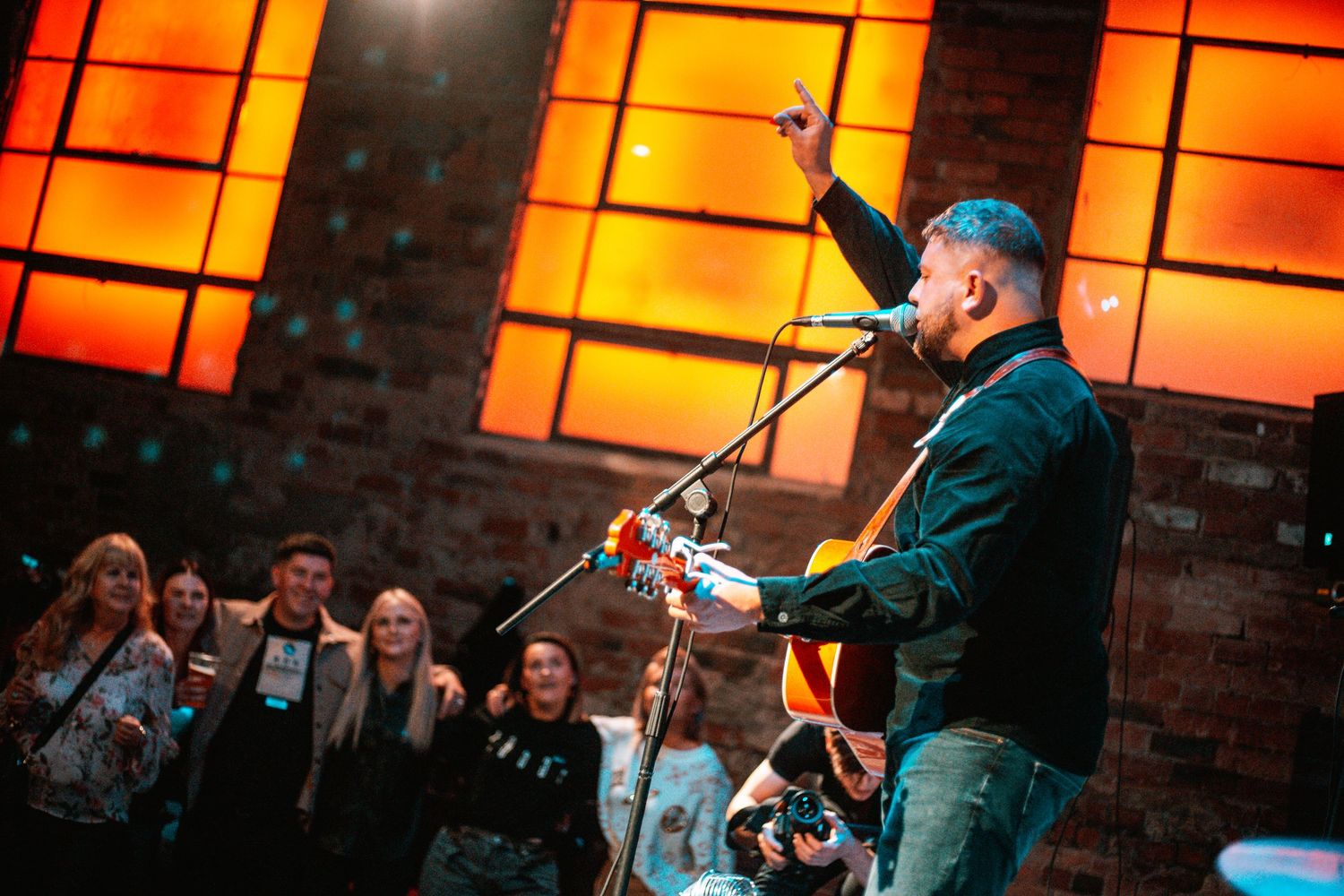 Musician performs with guitar against orange-lit windows as crowd watches.