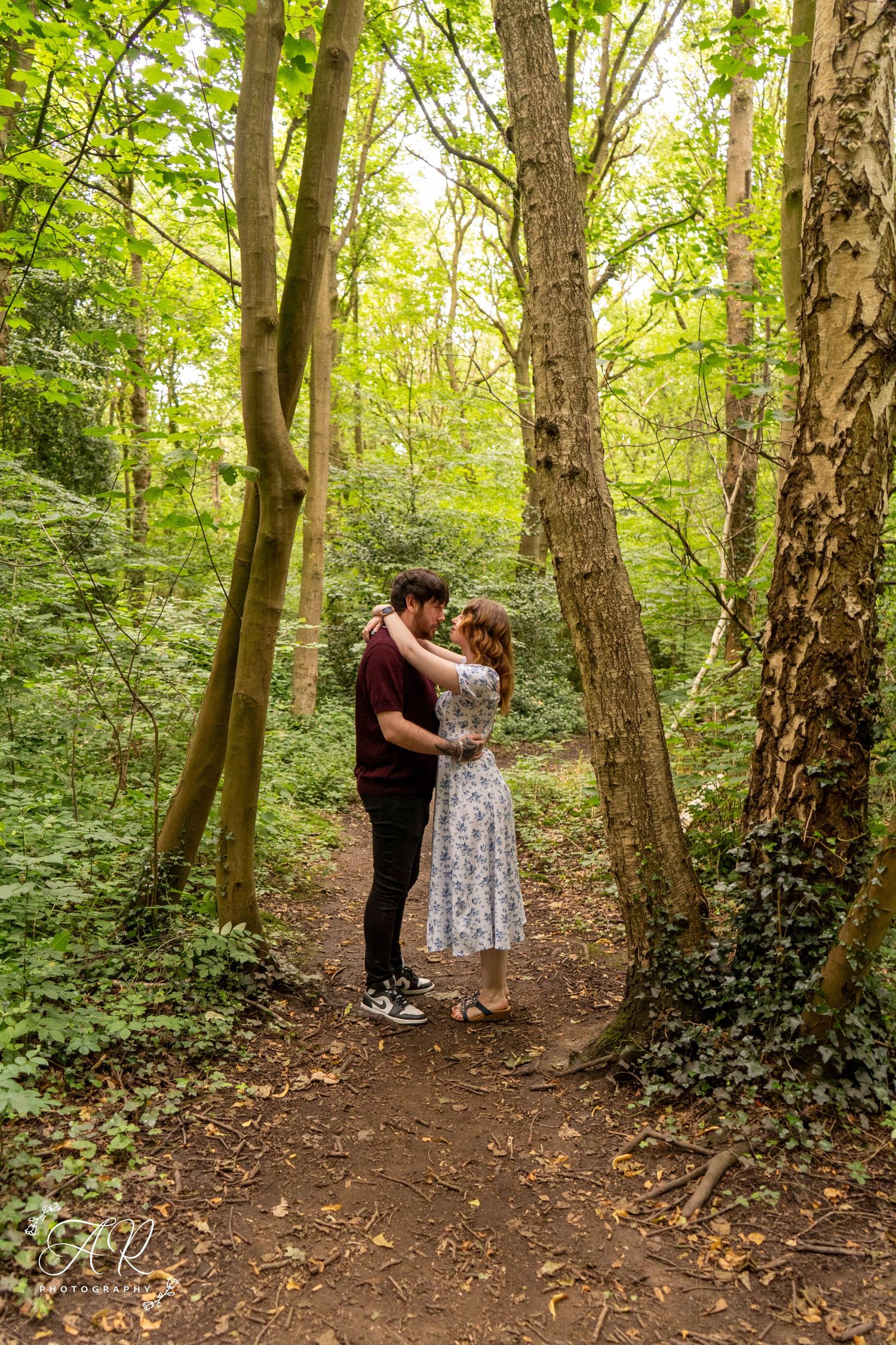 A couple embraces on a forest path framed by tall trees and ivy.