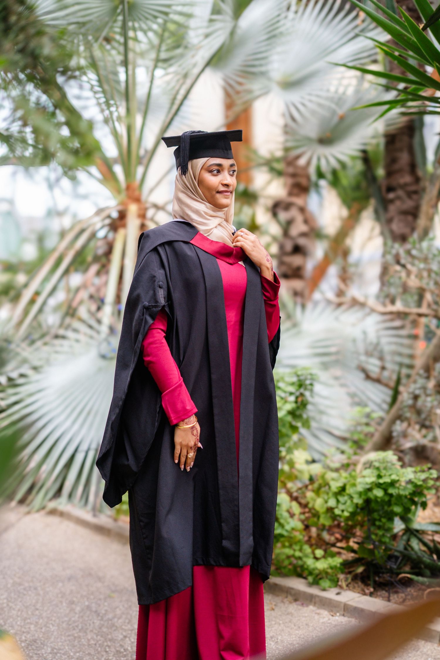 Graduate poses in academic dress among palm trees in greenhouse environment.