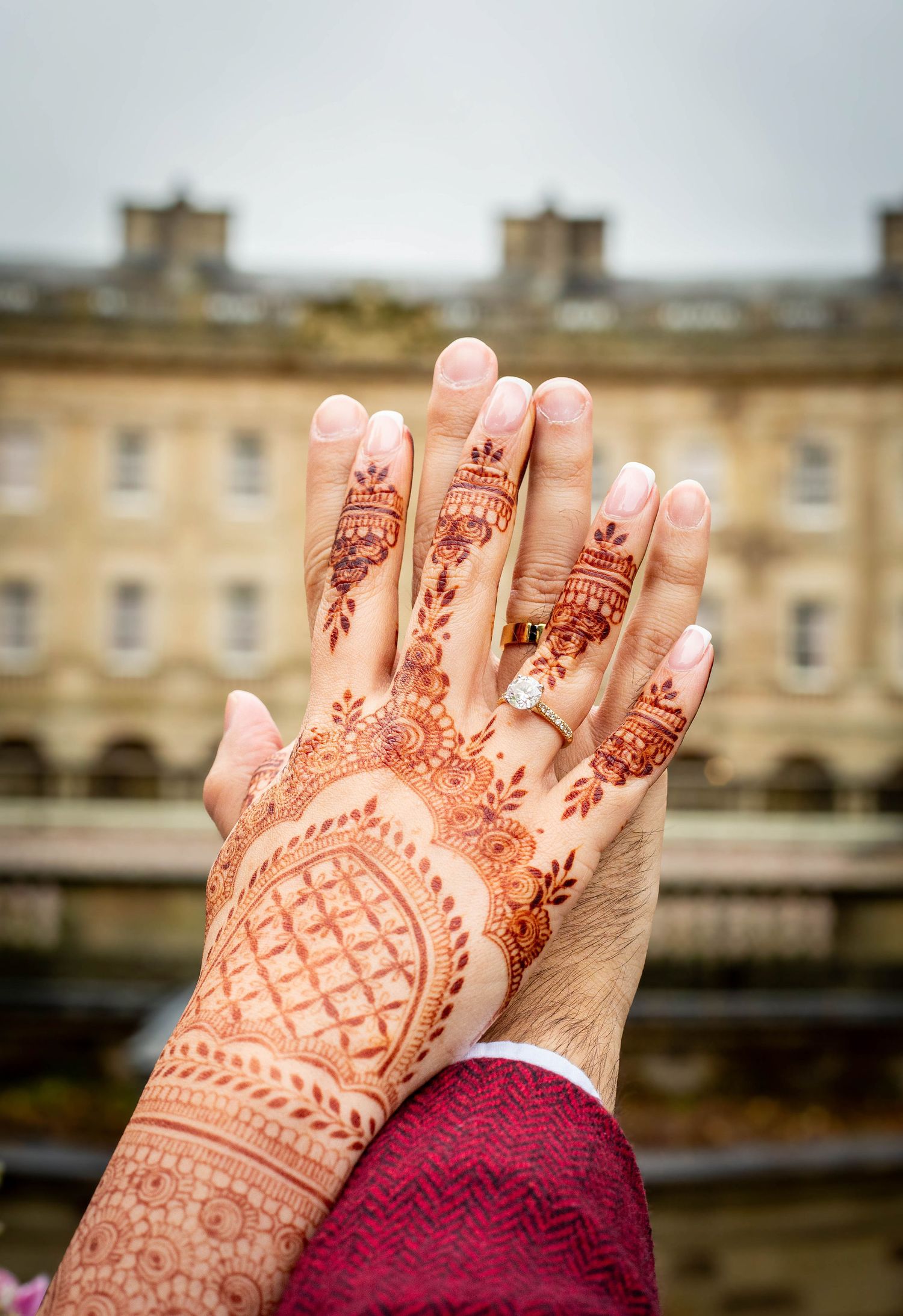 Detailed henna designs adorn hands with burgundy sleeves against historic building backdrop.