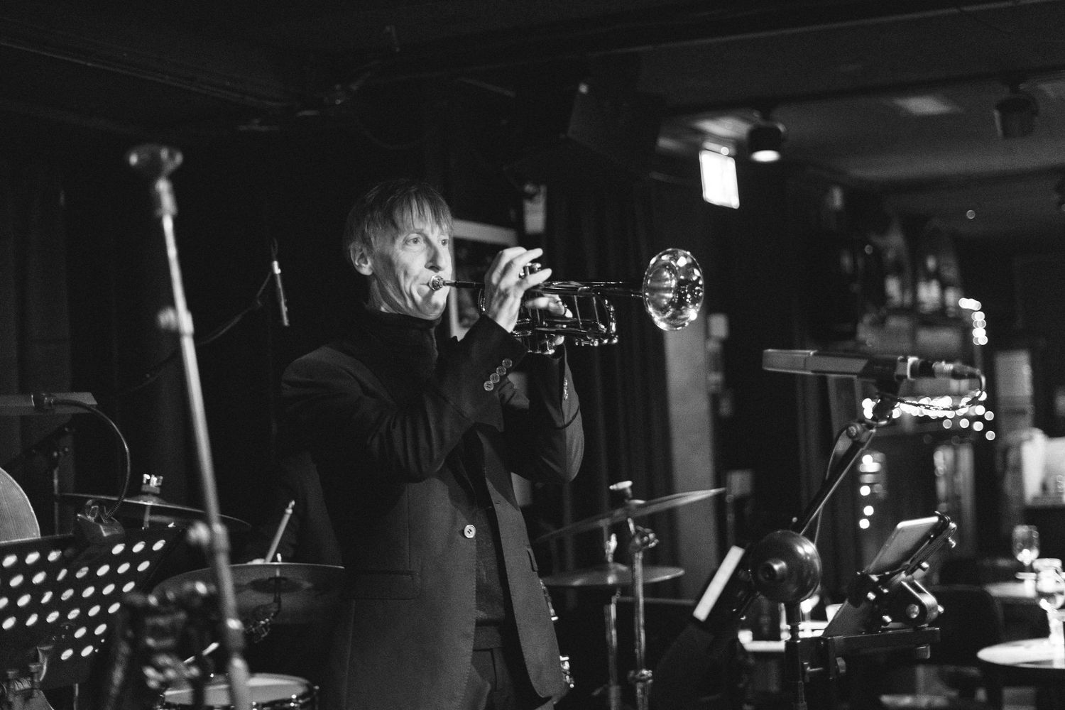 A black and white photo of a trumpet player performing on stage with a microphone stand visible.
