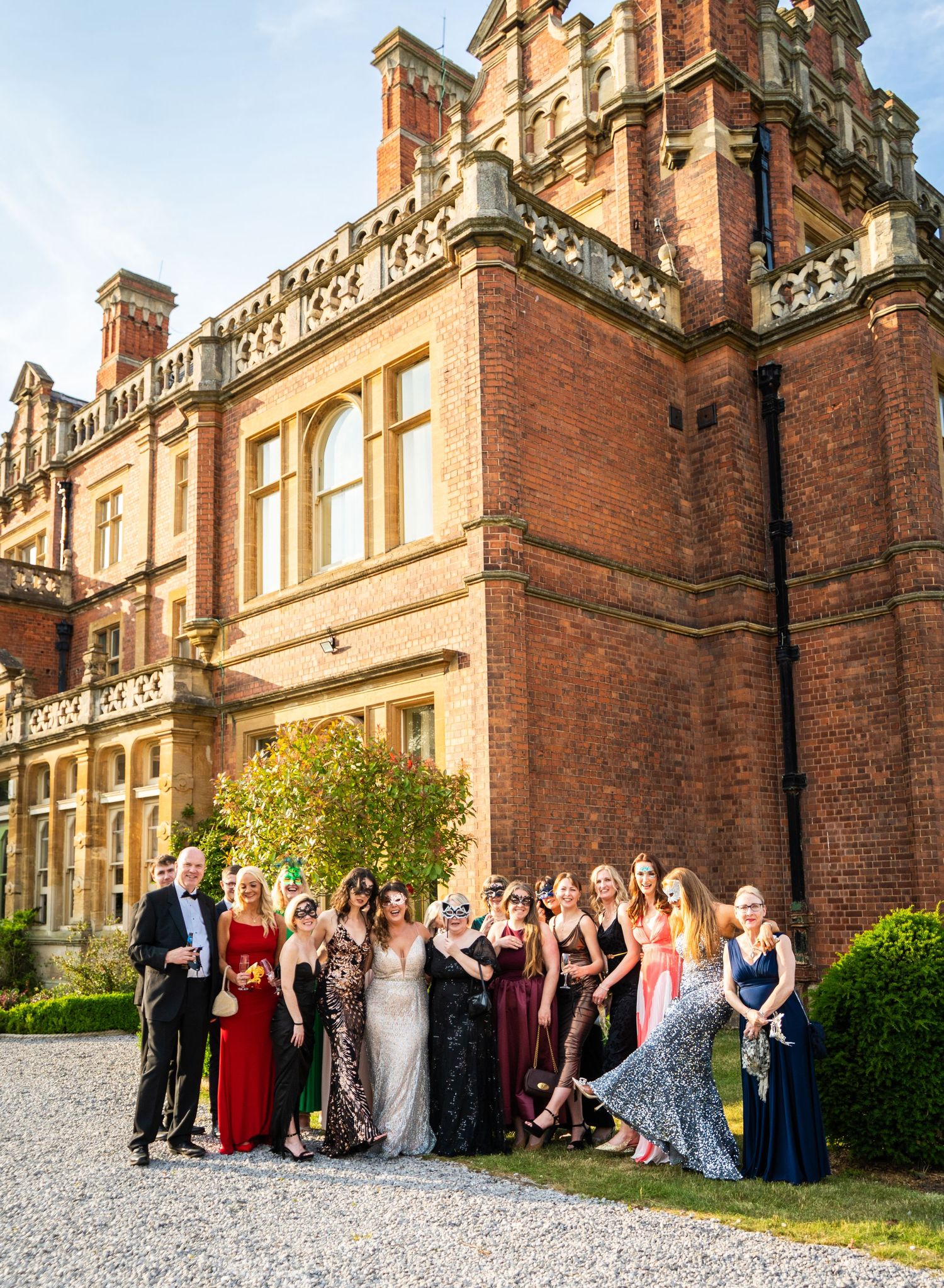 Formal group photo of elegantly dressed guests gathered outside a grand brick mansion for a special event.