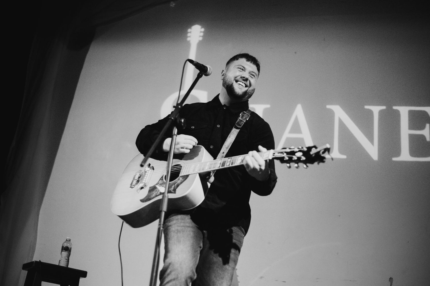 Musician performs on acoustic guitar in black and white against illuminated backdrop with text.