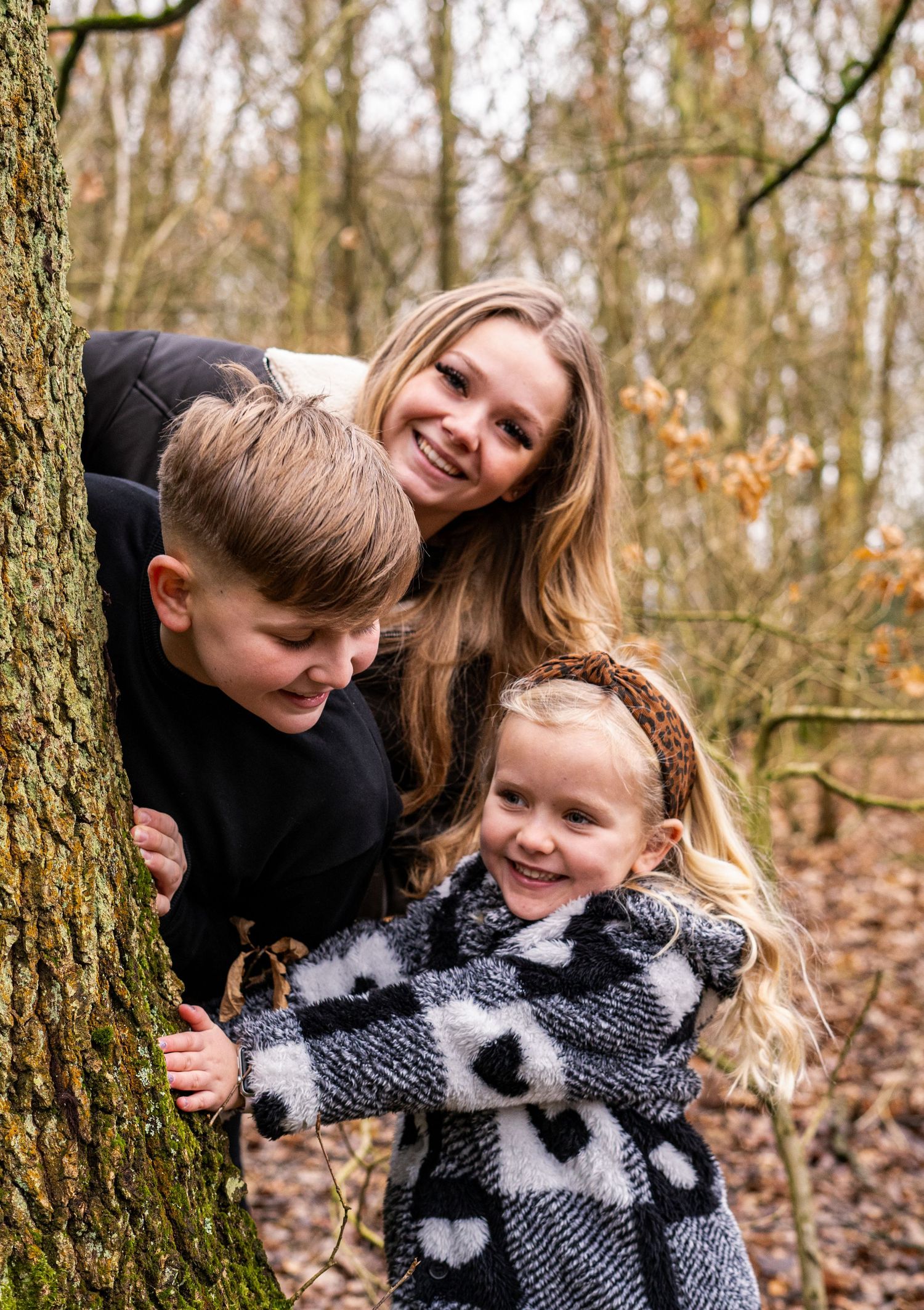A playful family photo session in an autumn forest with people peeking around a large tree trunk.