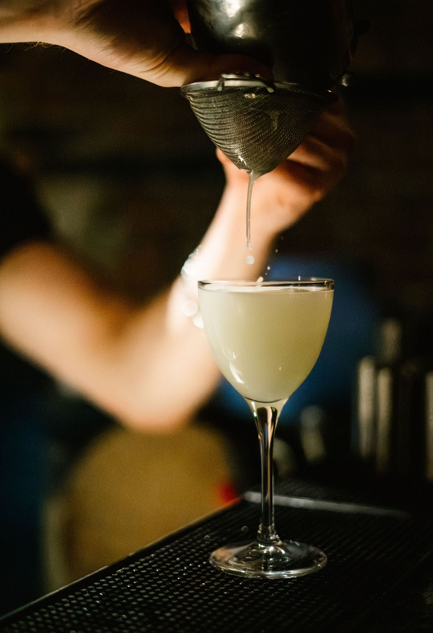 A creamy cocktail being poured into an elegant coupe glass at a dark bar.