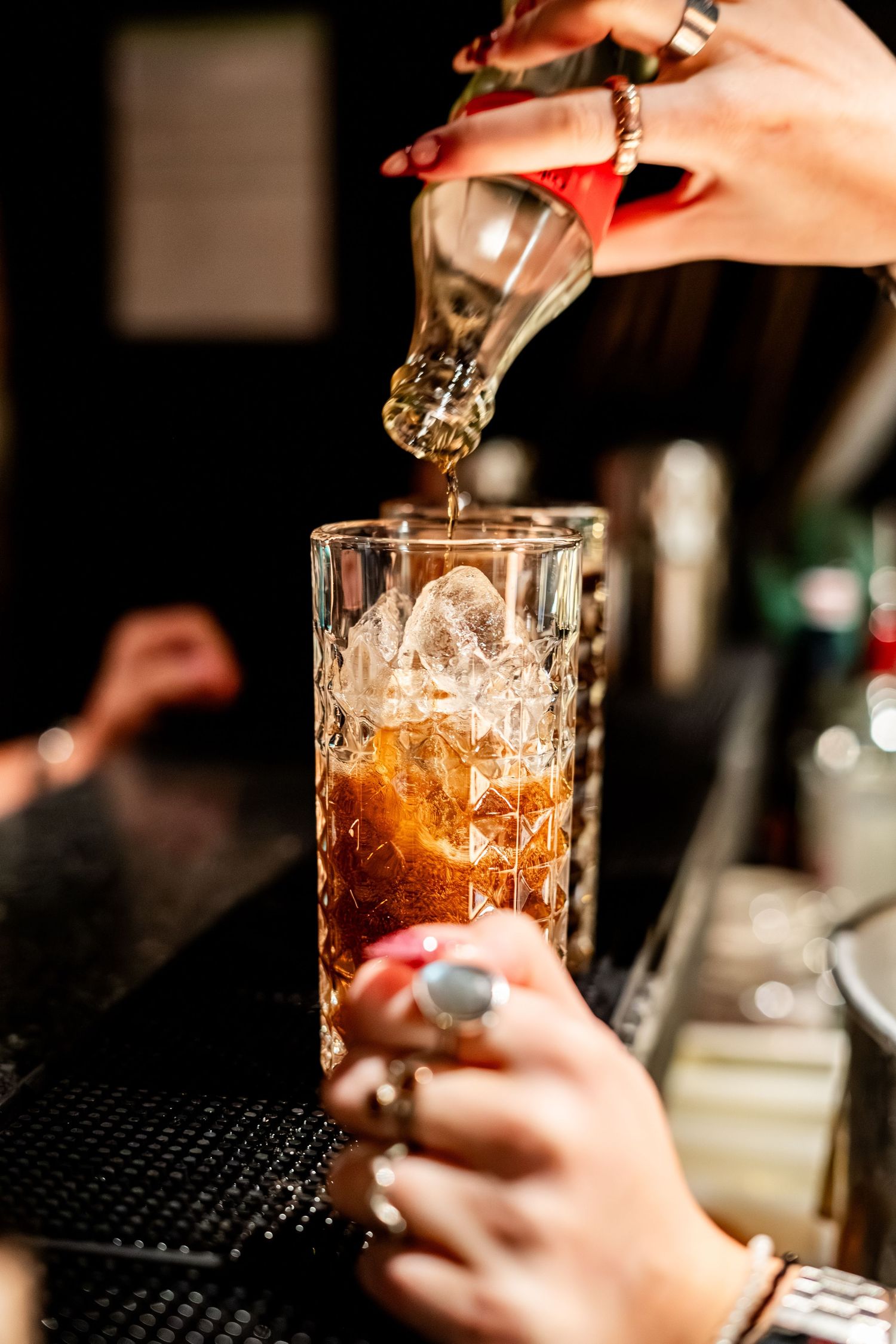 Hands pouring amber liquid into a crystal mixing glass with ice.